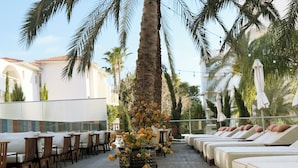A rooftop lounge with lounge chairs and tables, surrounded by palm trees and a decorative flower arrangement.