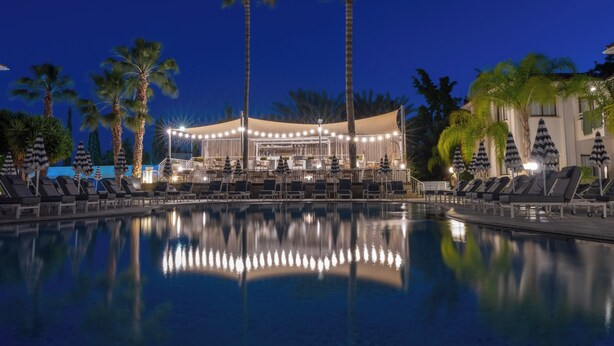 A nighttime pool area with lounge chairs and umbrellas, palm trees, and a building with string lights.