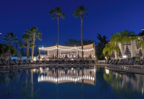 A nighttime pool area with lounge chairs and umbrellas, palm trees, and a building with string lights. - King Jason Paphos - Designed for Adults By Louis Hotels (Paphos)