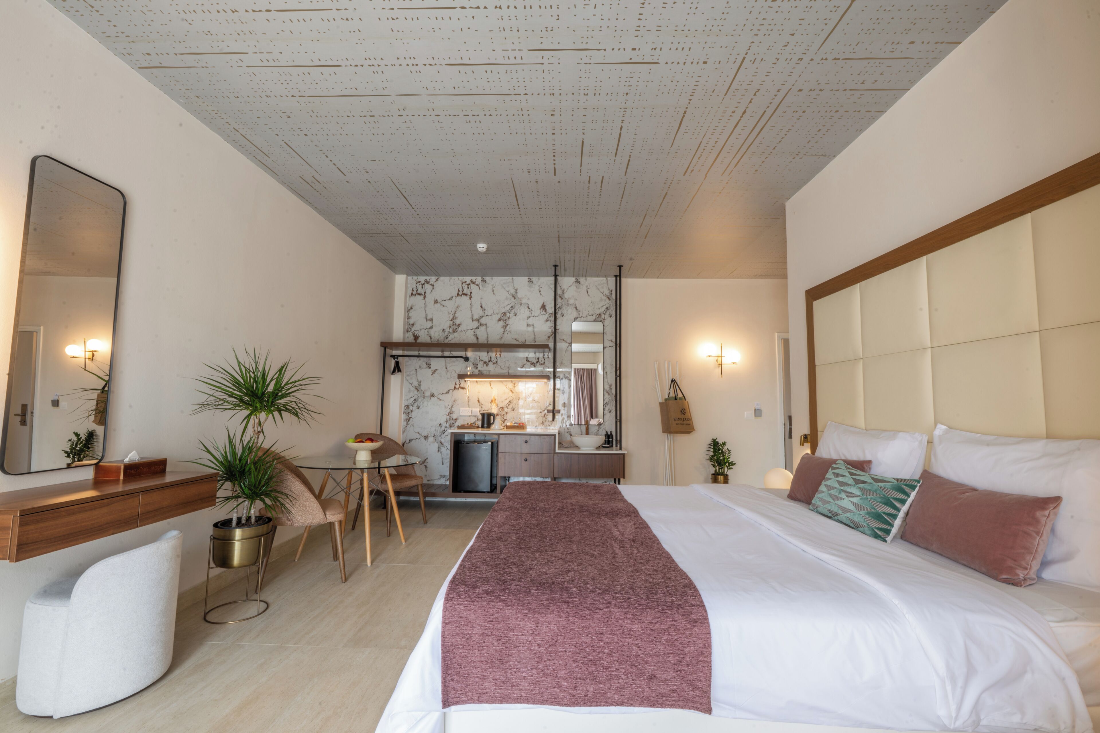 A modern hotel room with a bed, bedside table, chair, desk, and a kitchenette area.
