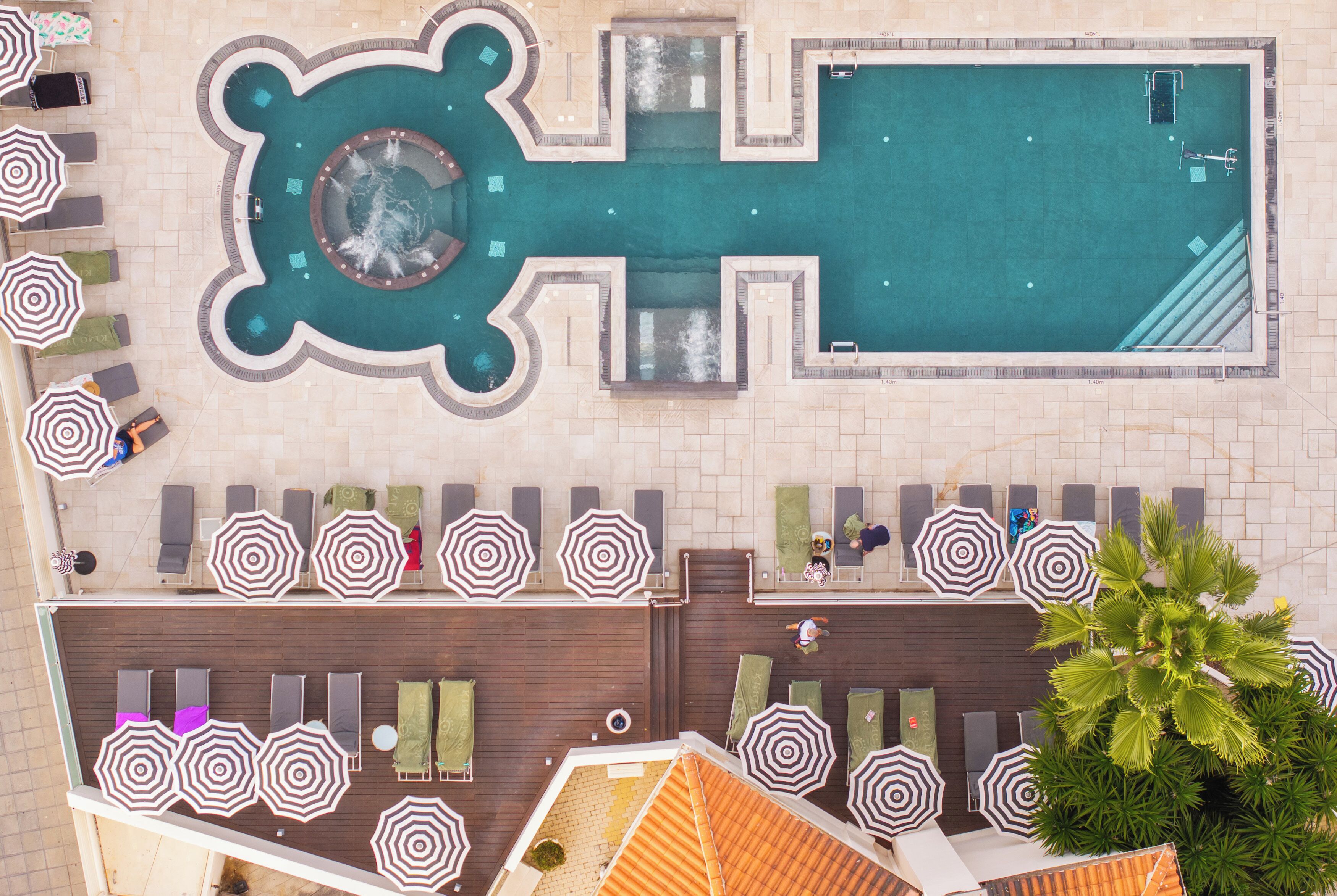 An aerial view of a hotel pool area with a central hot tub, surrounding lounge chairs, and umbrellas.