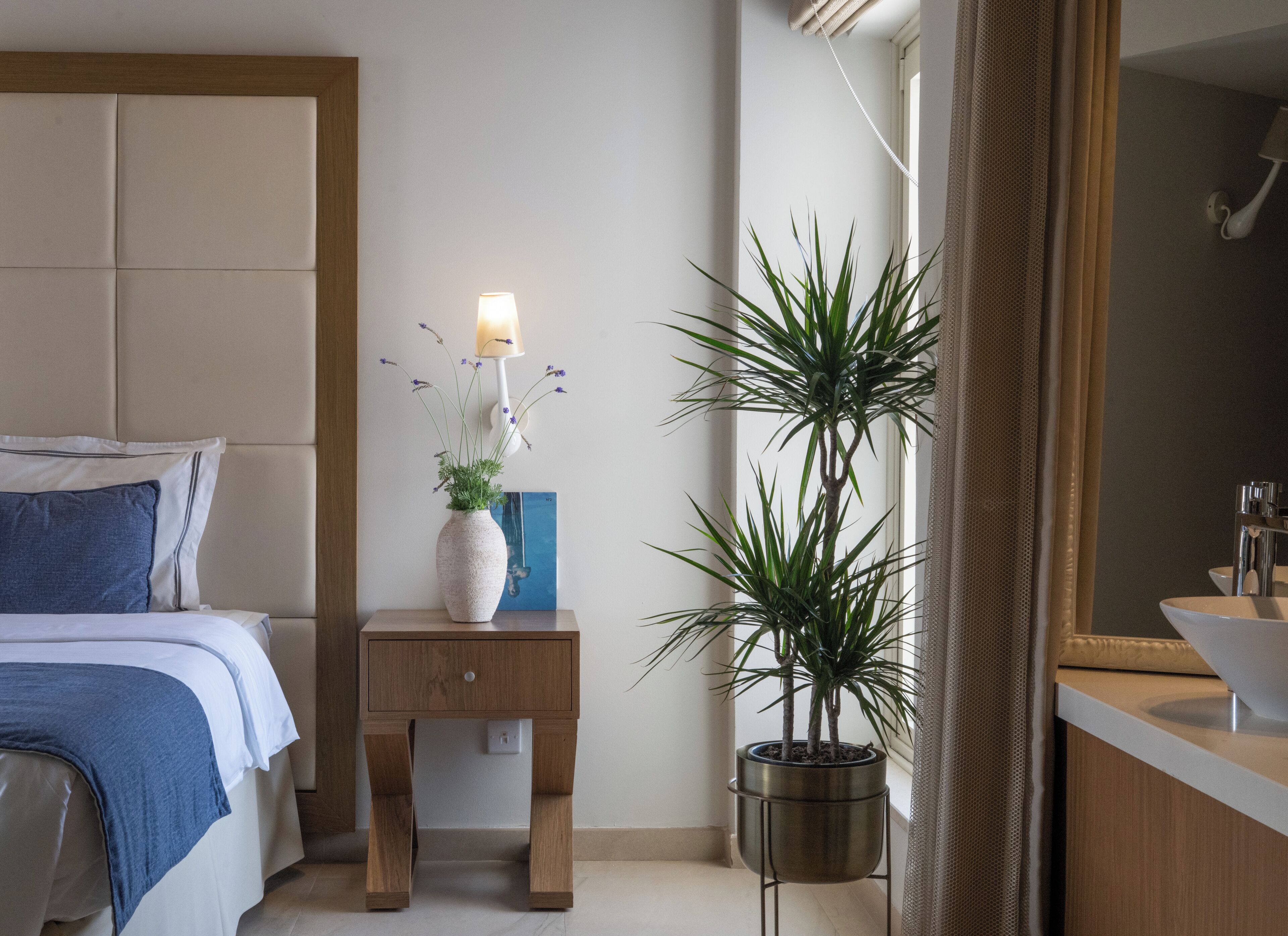 Een slaapkamer met een bed, nachtkastje met lamp, een potplant en een badkamer met wastafel en spiegel.