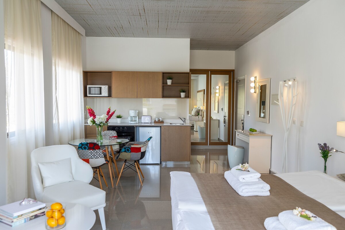 Habitación de hotel con cama, escritorio, silla, kitchenette pequeña y baño con espejo e iluminación.