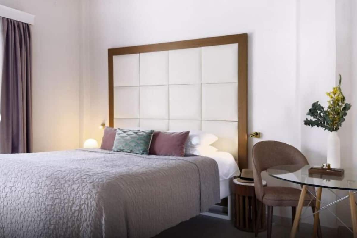 A hotel room with a large bed, a glass table, a chair, and a vase with flowers.