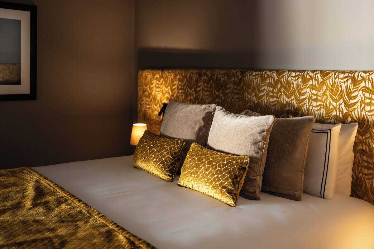 Uma cama bem arrumada com cabeceira estampada, abajur e um quadro na parede.