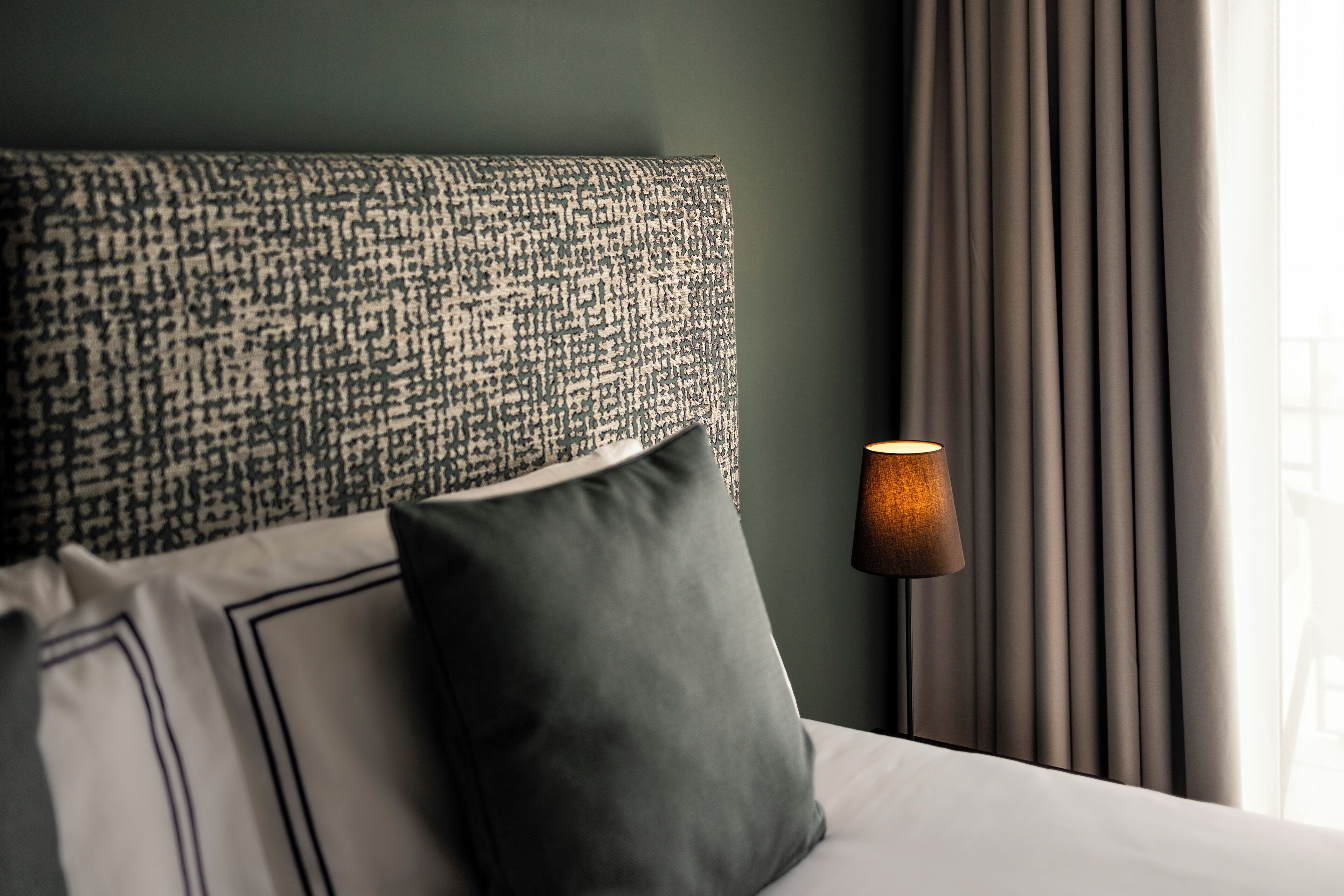 A neatly made bed with a patterned headboard, a dark pillow, and a lamp on a bedside table.