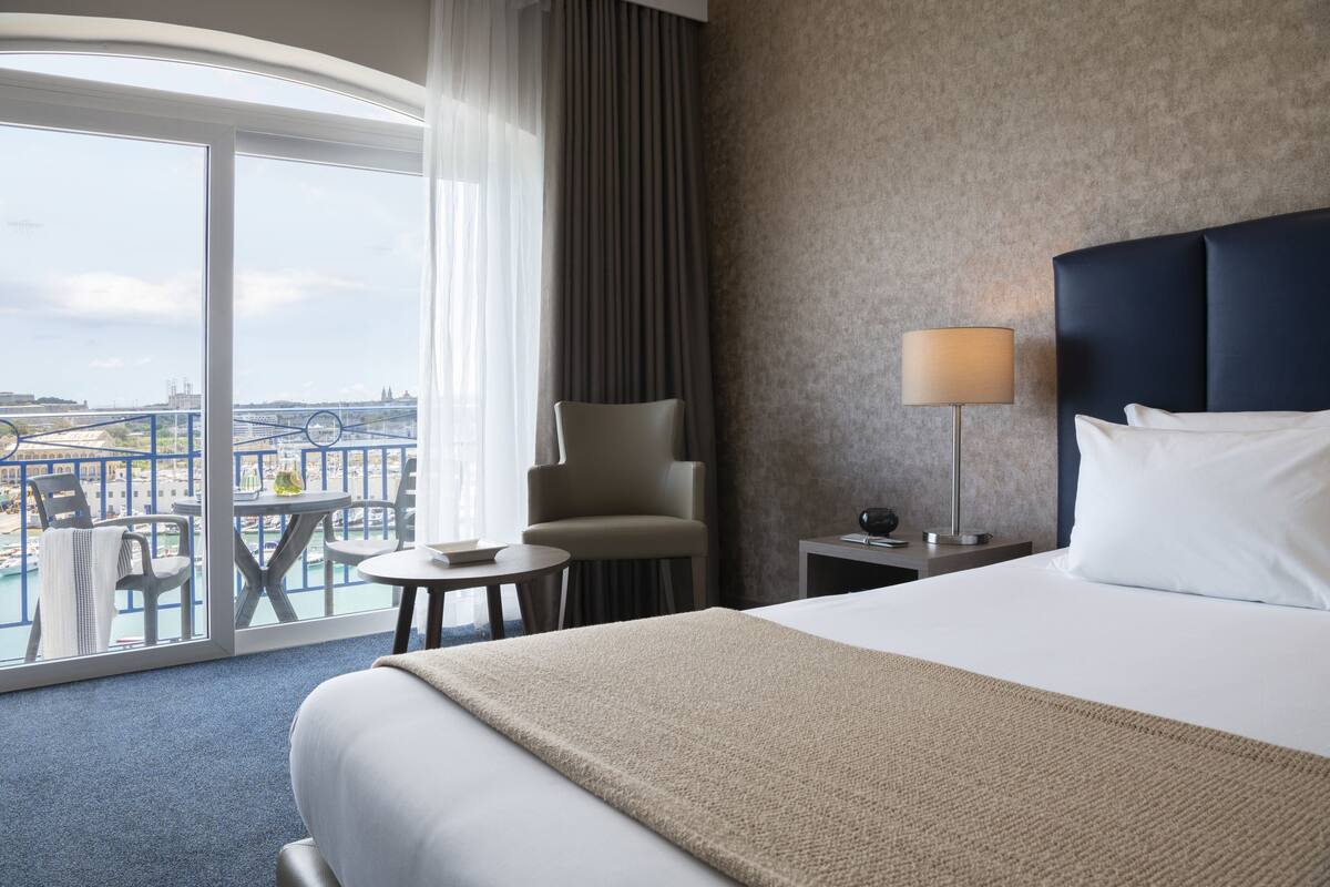 Quarto de hotel com uma cama grande, uma cadeira, uma mesa pequena, um abajur e vista para a paisagem urbana através de uma varanda.
