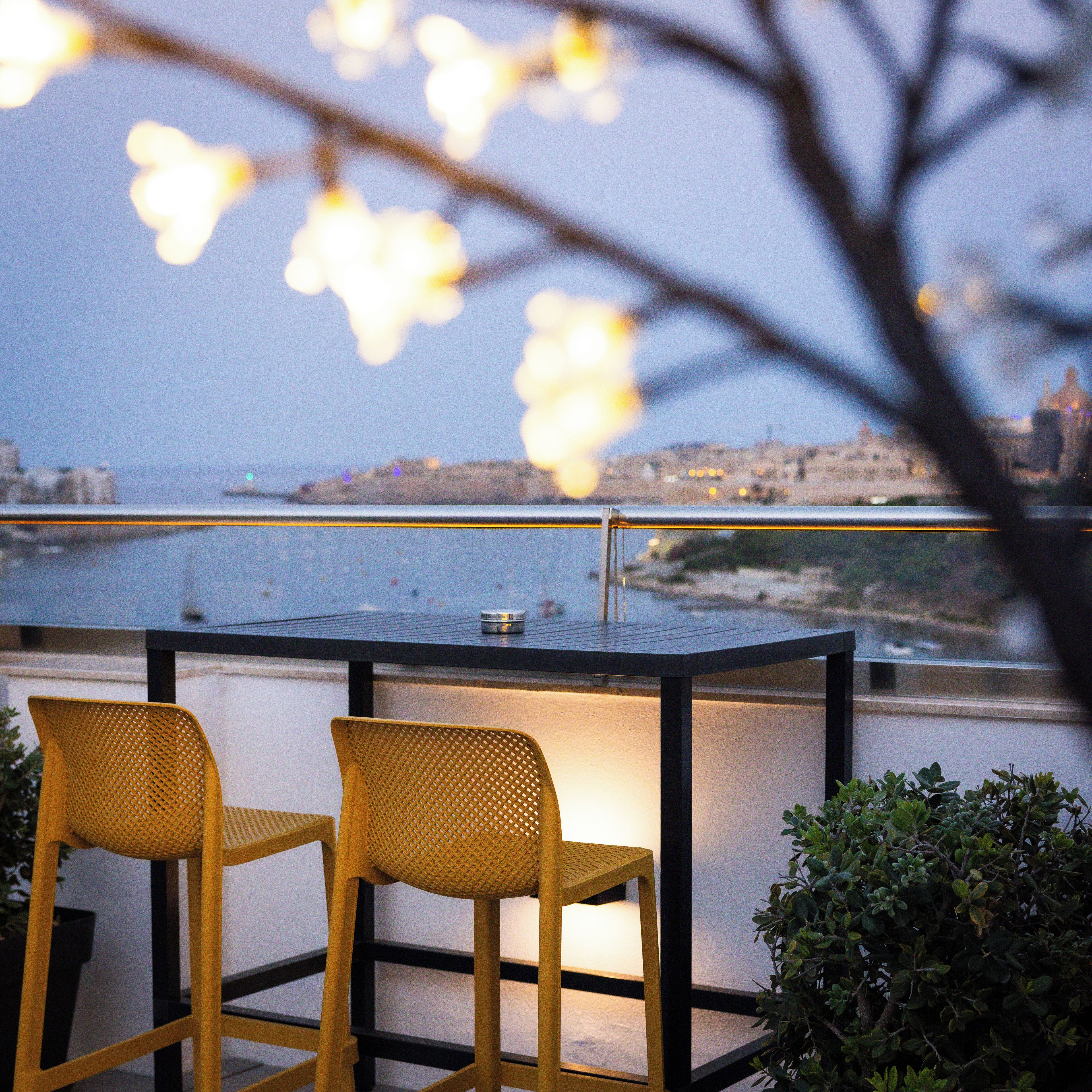 Un balcon avec deux chaises jaunes et une table, d’où l’on peut admirer un paysage urbain et une étendue d’eau.