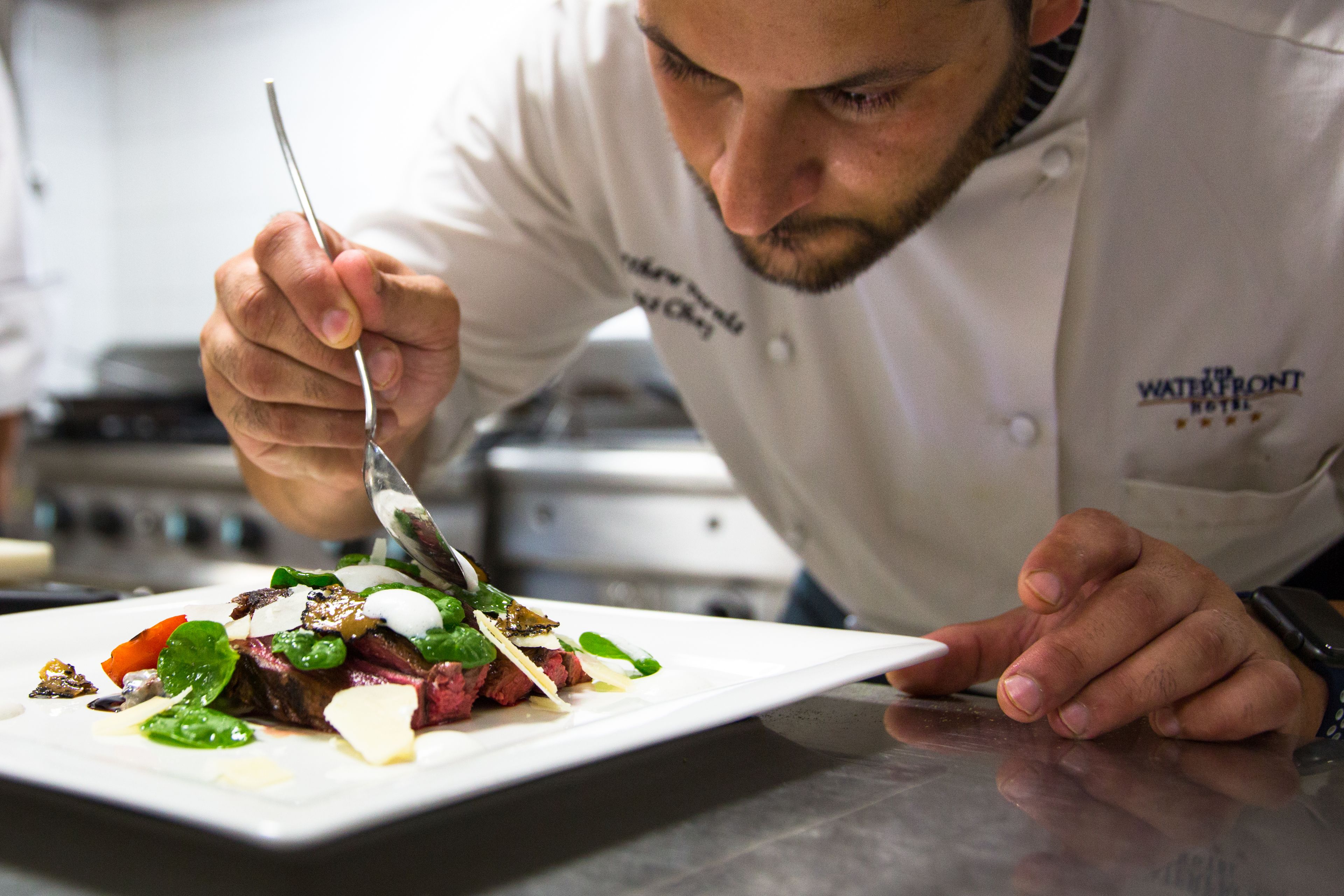 A chef in a white uniform with a logo, preparing a dish on a white plate.
