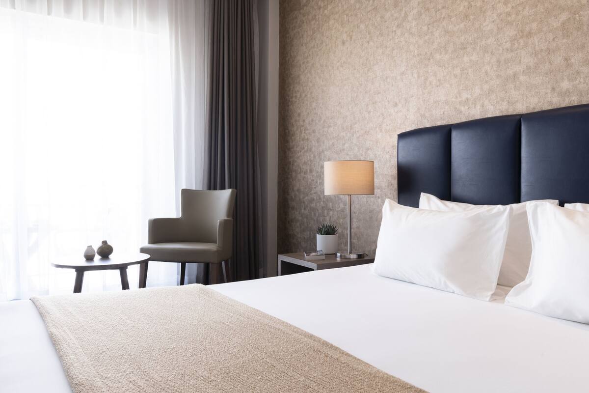 Quarto de hotel com uma cama grande, uma cadeira, uma mesinha e um abajur.