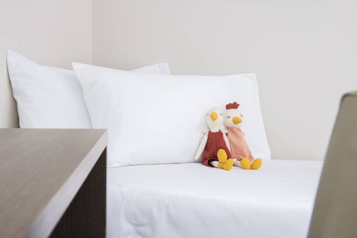 Cama bem arrumada com roupa de cama branca e um brinquedo de frango macio sobre o travesseiro.