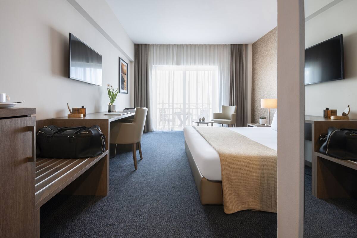 Quarto de hotel com cama, escrivaninha, cadeira e televisão fixada na parede.