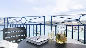 Un balcón con vistas a la ciudad y al agua, una mesa con un libro, vasos y una jarra de vidrio.