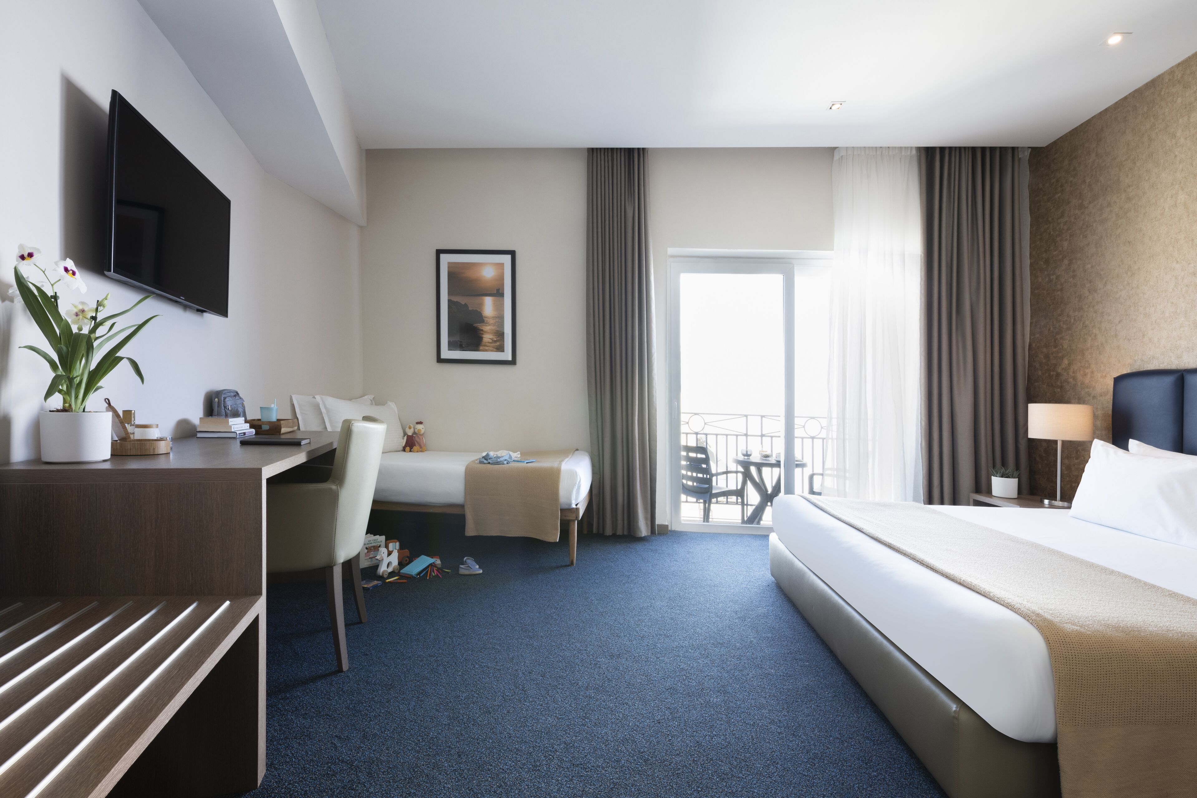Quarto de hotel com cama, escrivaninha, cadeira, televisão, janela com cortinas e varanda.