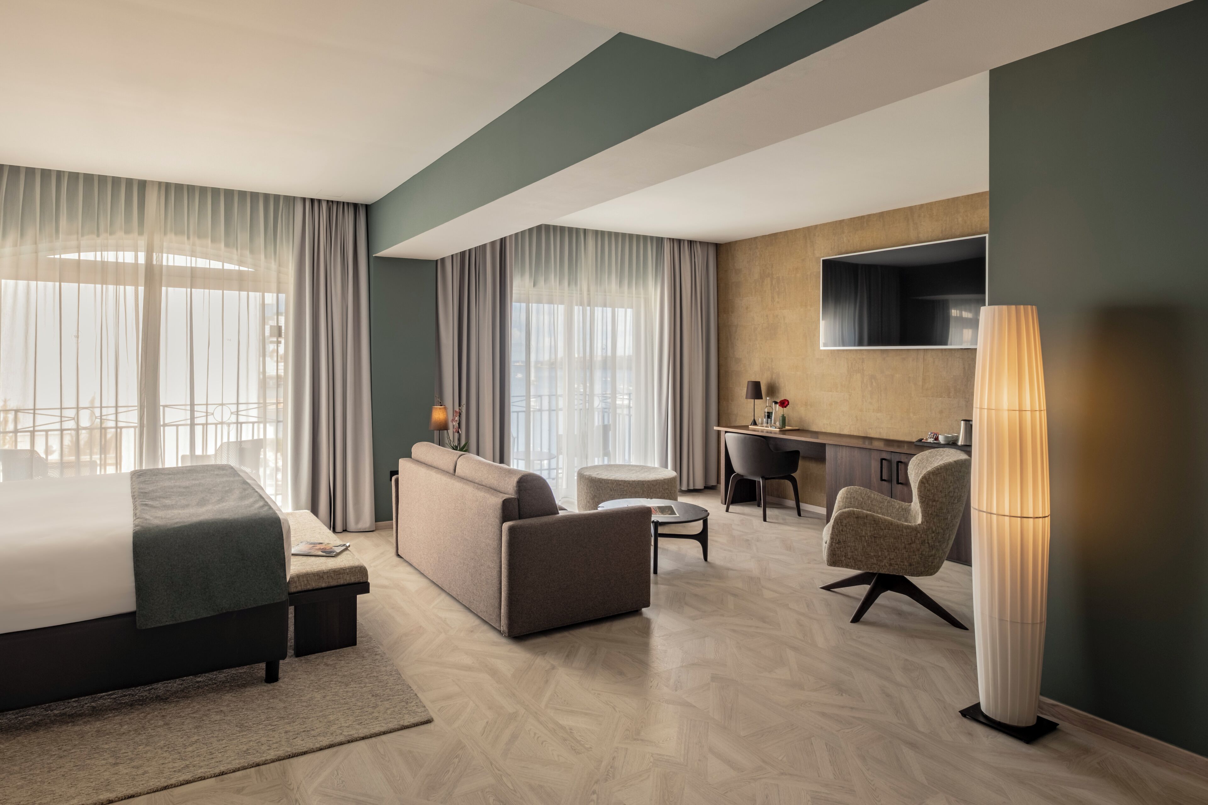 A modern hotel room with a bed, sofa, desk, and chair, featuring a large window and a TV.