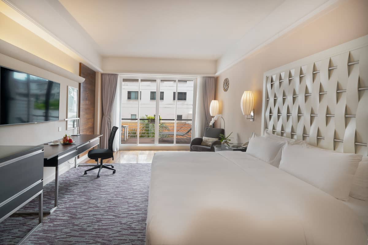 Habitación de hotel con una cama grande, un escritorio con silla, televisión, lámpara y vistas al balcón exterior.