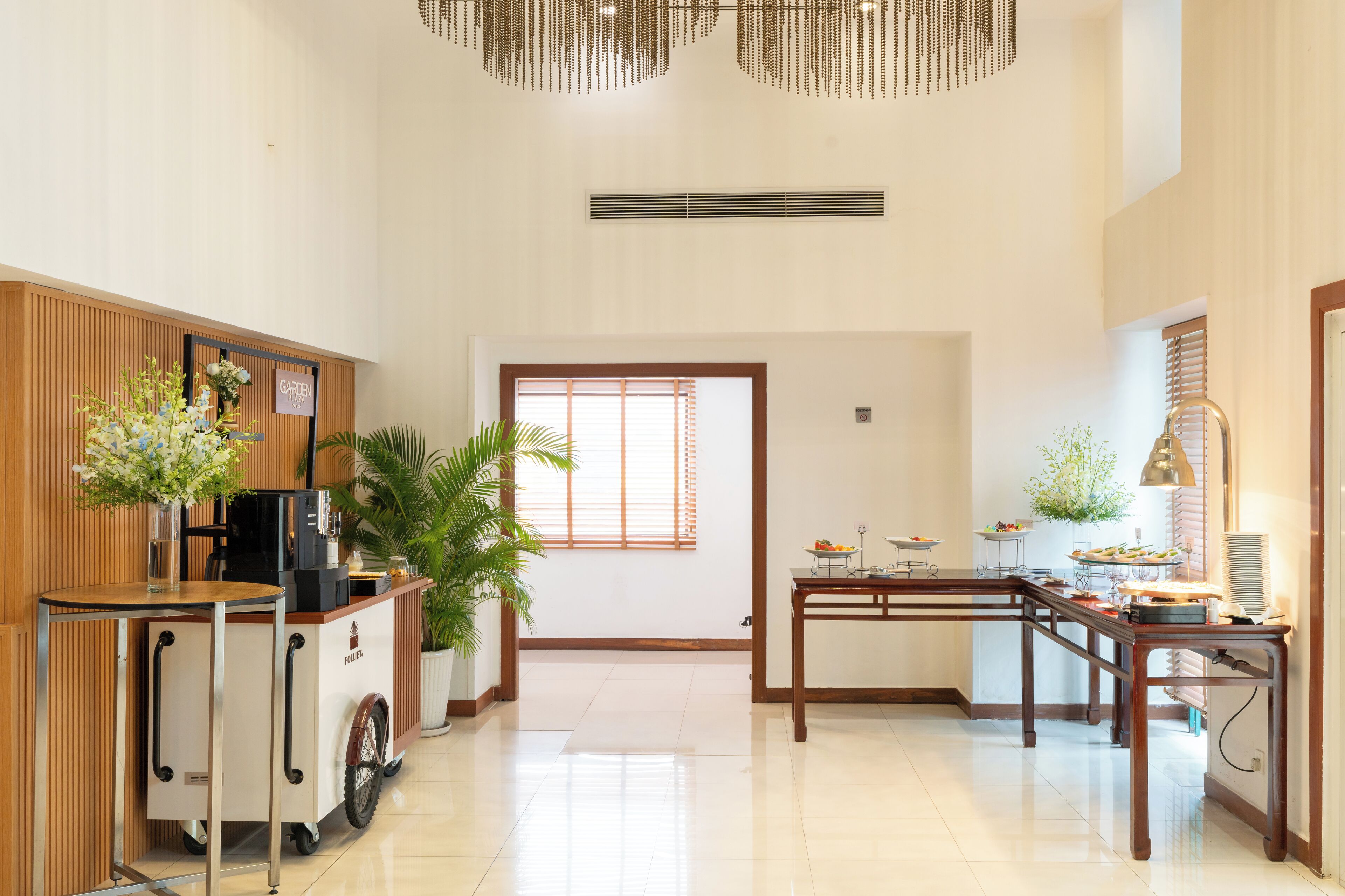 A hotel lobby with a coffee station, a reception desk, a dining area, and a large window with blinds.