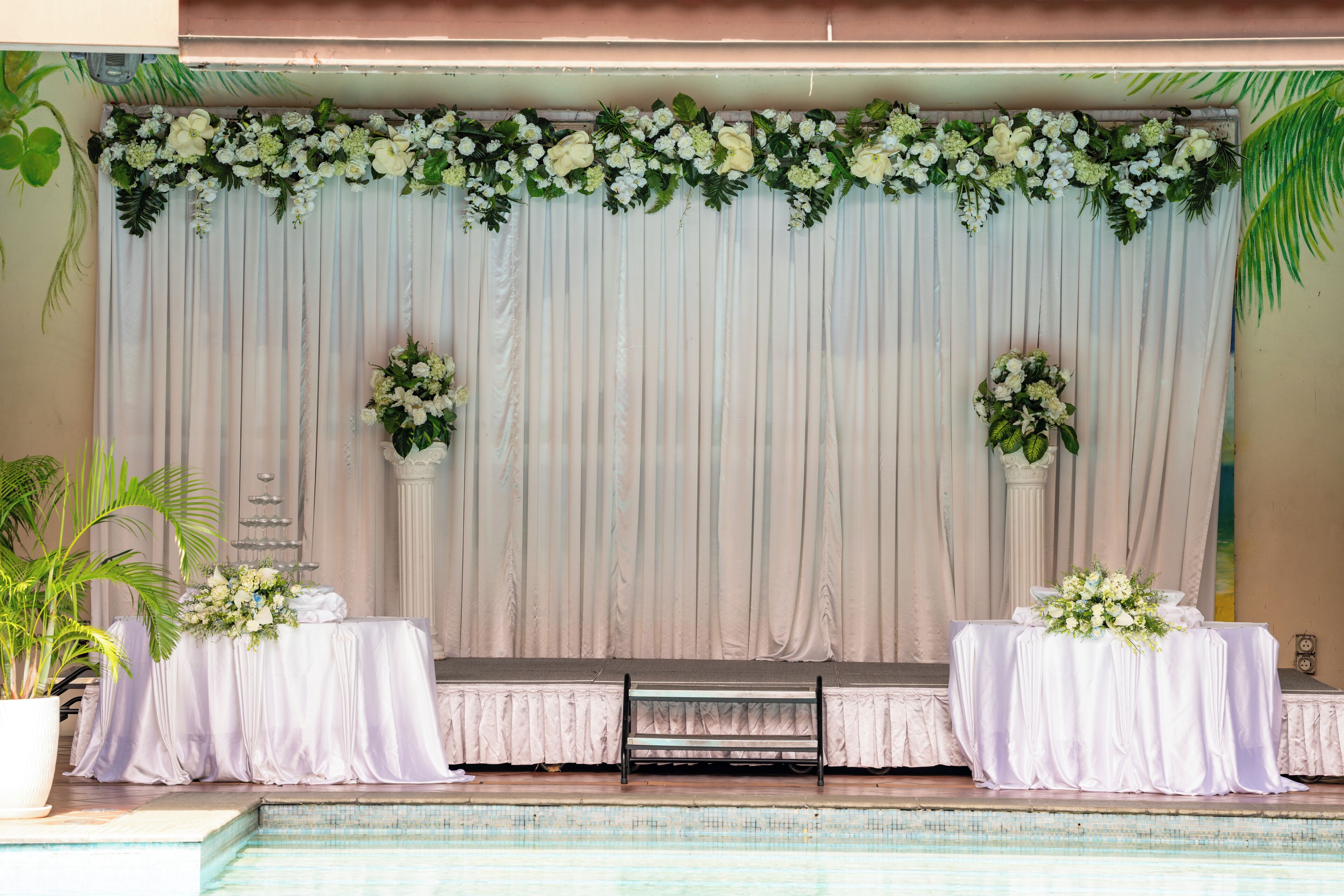 Un escenario con un telón blanco de fondo, dos mesas laterales con arreglos florales y un banco en el centro.
