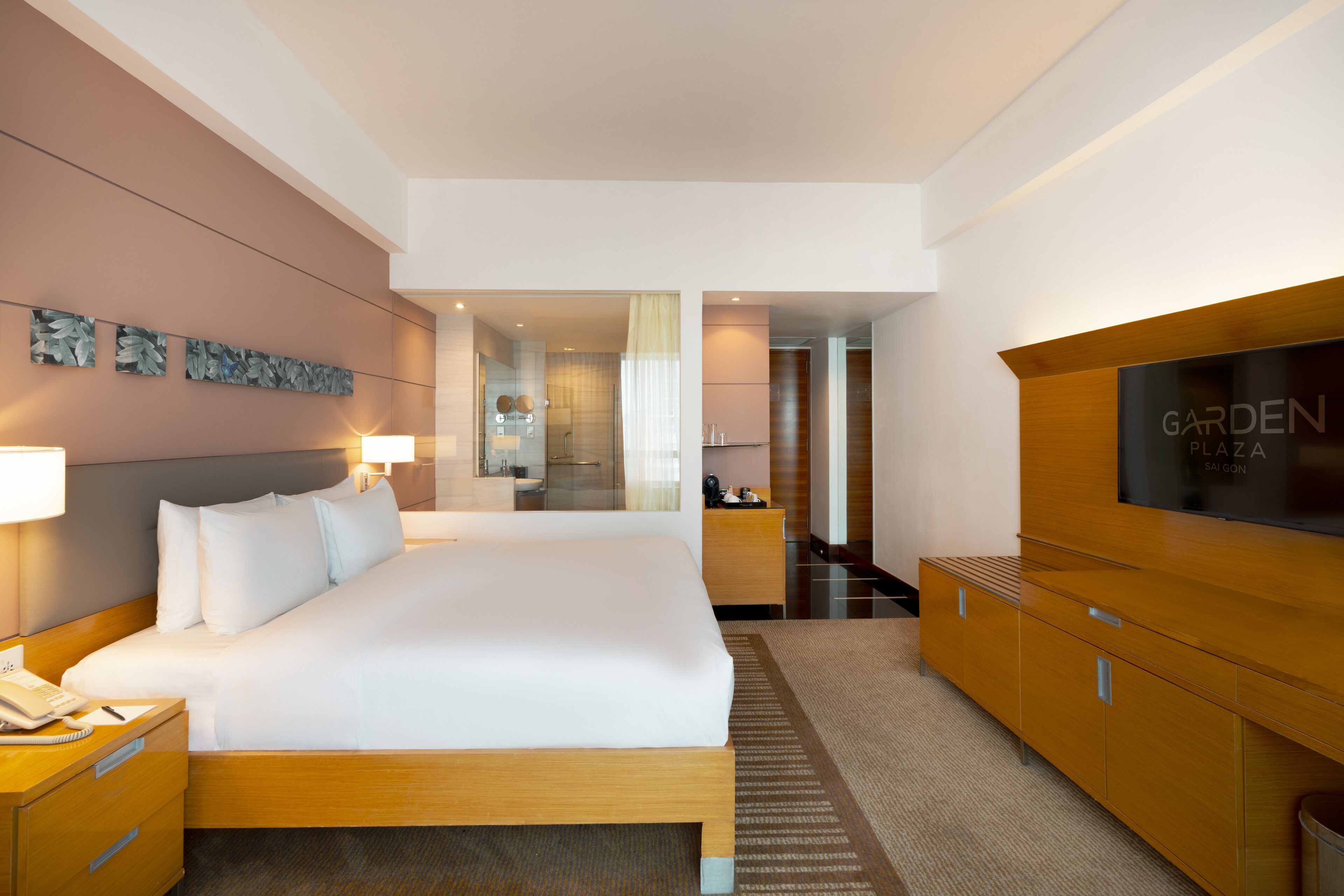 A hotel room with a large bed, wooden headboard, a TV on a wooden cabinet, and a bathroom visible through a glass door.
