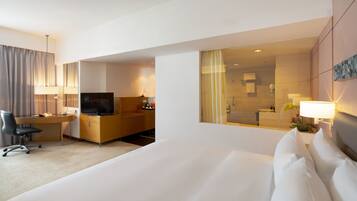 A hotel room with a large bed, a desk with a computer, a TV, and a bathroom visible through a glass partition.