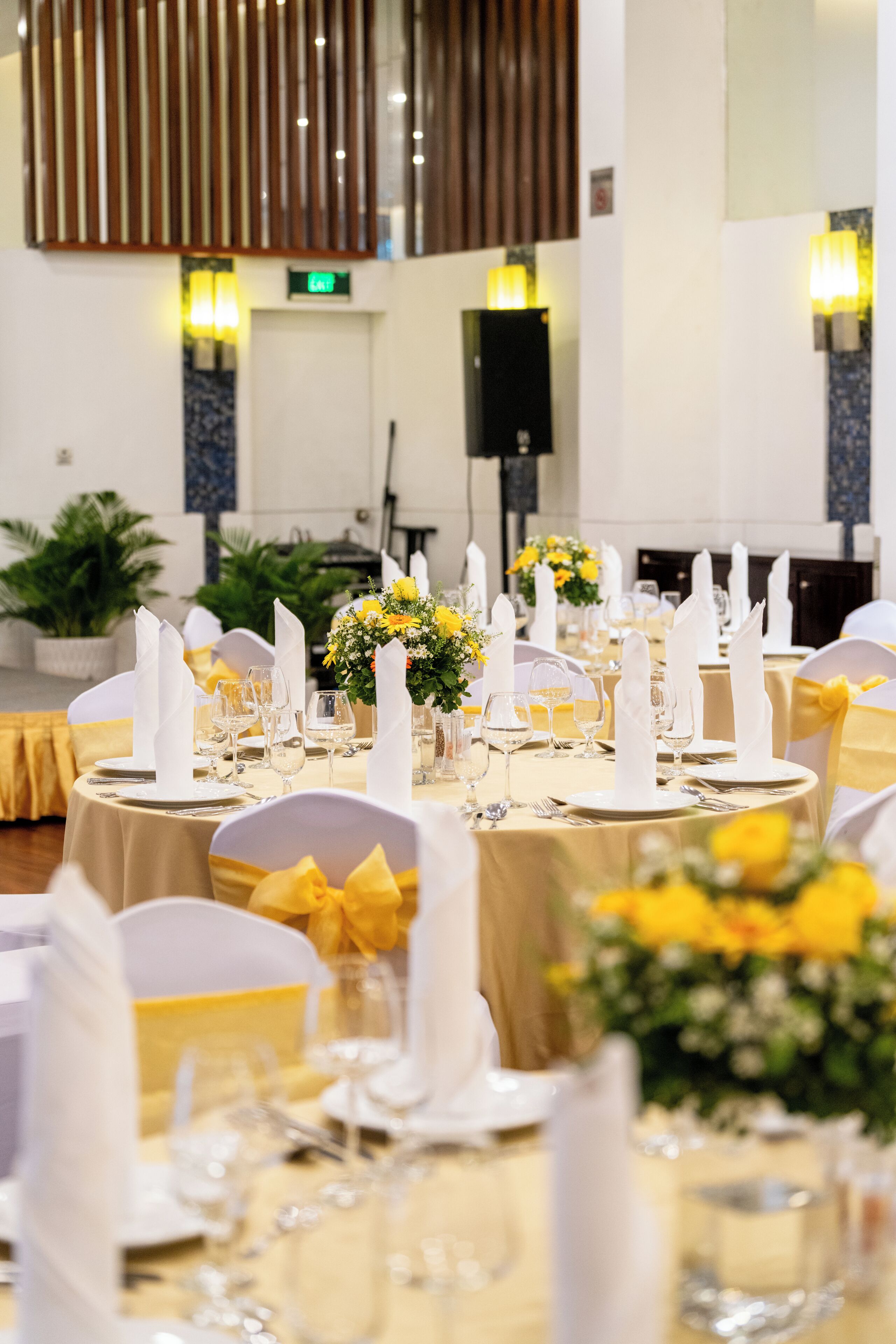 A banquet hall with round tables set for a formal event, featuring white napkins, glassware, and yellow floral centerpieces.