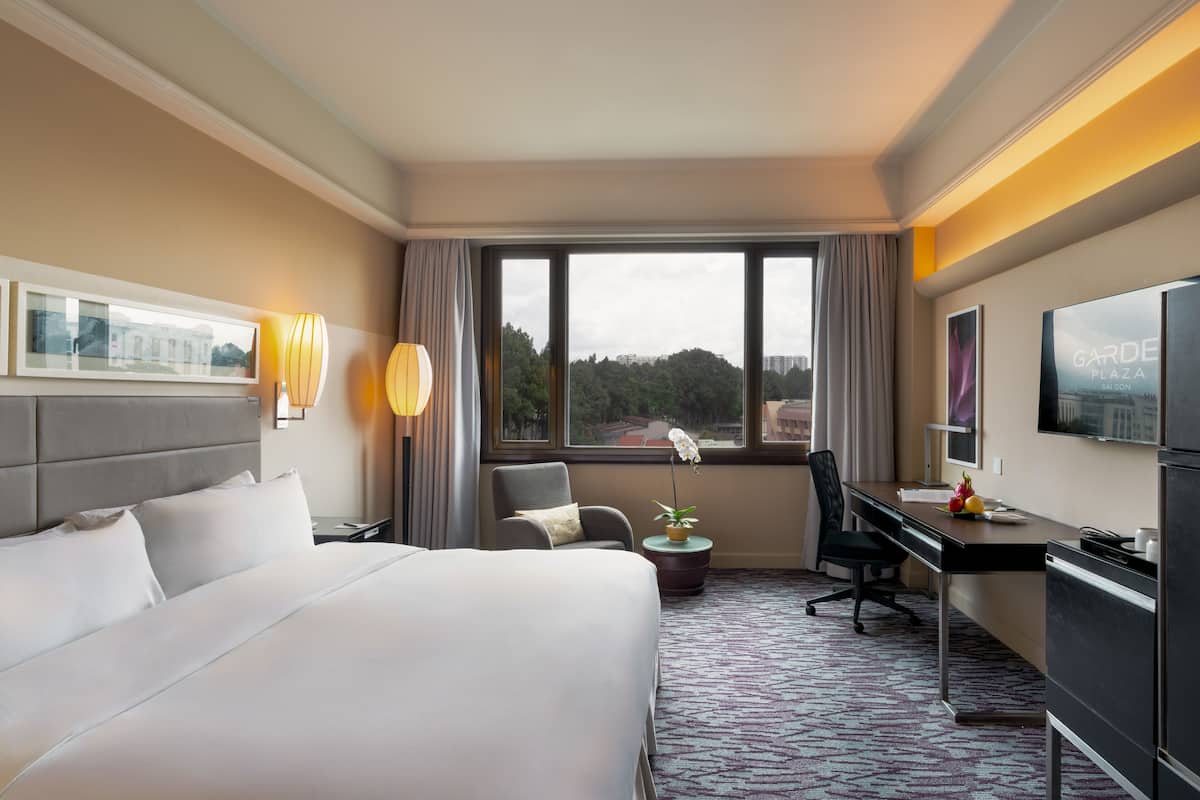 Habitación de hotel con una cama grande, un escritorio, una silla, un televisor y una ventana que da a vistas de edificios y árboles.