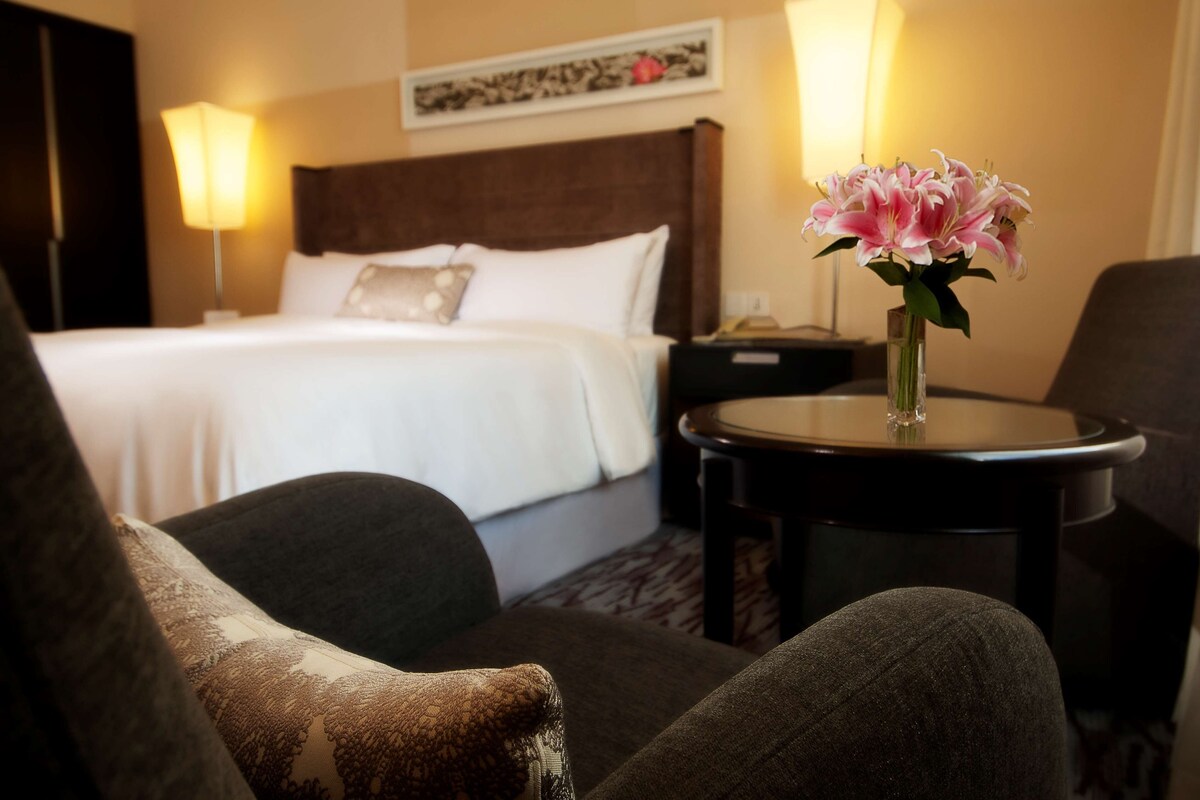 Une chambre d’hôtel avec un lit, une table de chevet, une lampe, un vase avec des fleurs et une chaise.