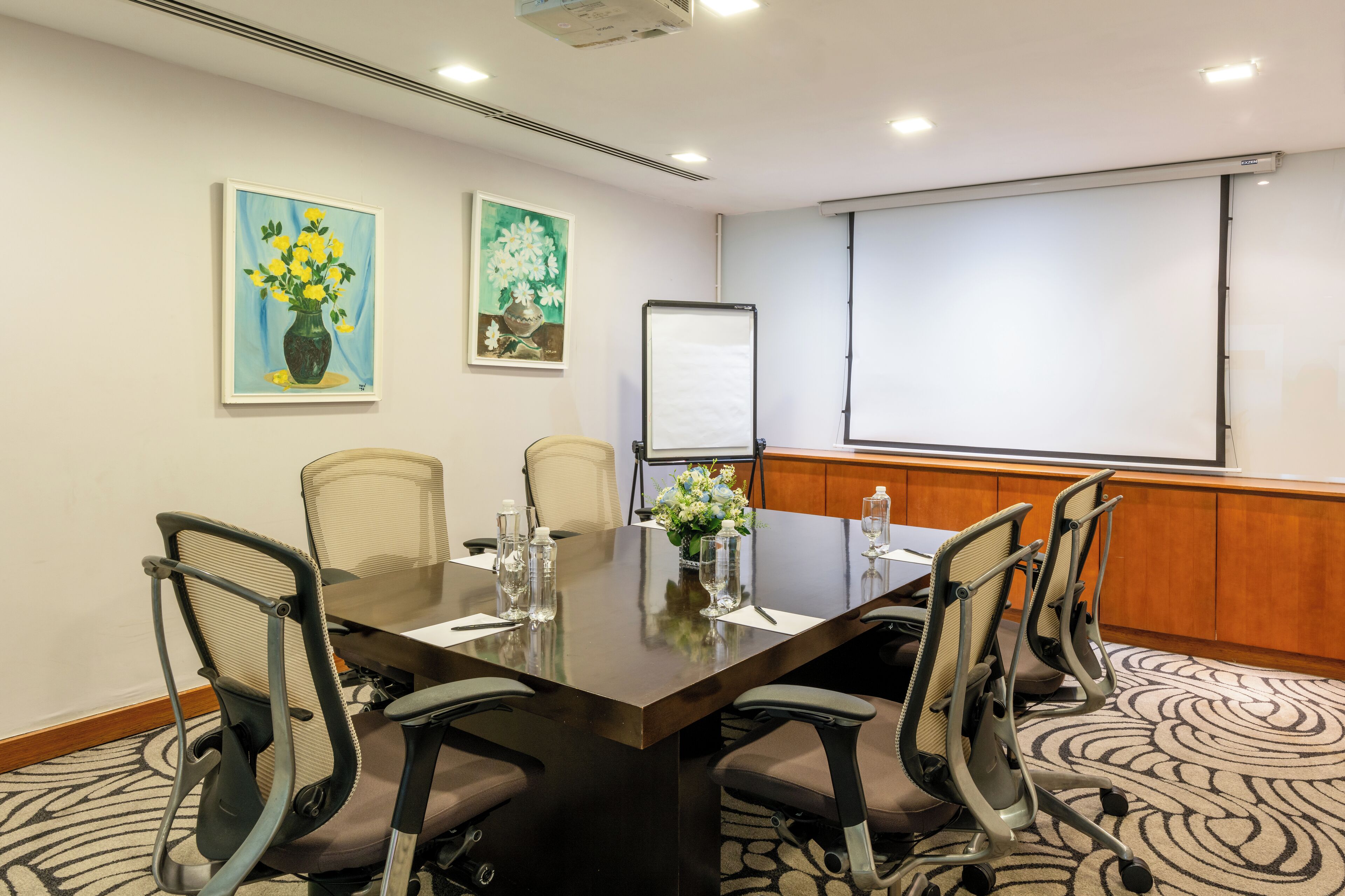 Un salón de conferencias con una mesa grande, seis sillas, una pantalla de proyector y dos cuadros en la pared.