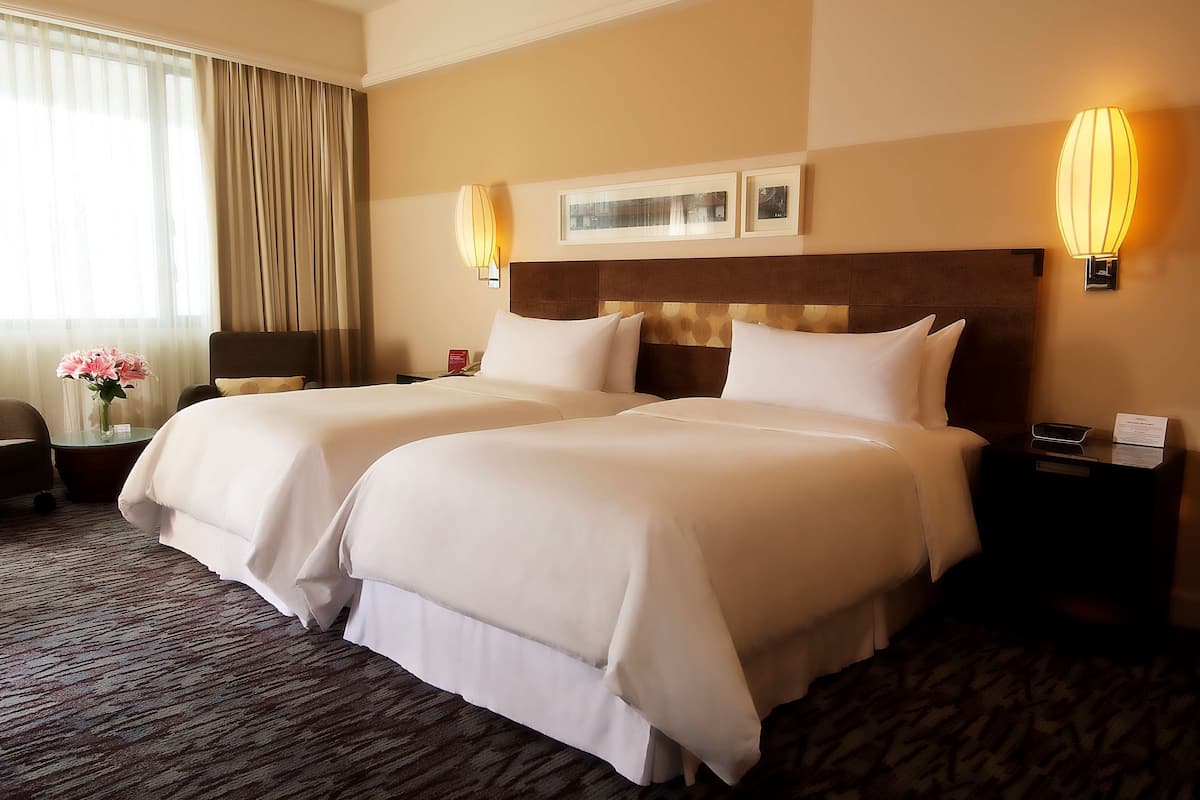 Habitación de hotel con dos camas, una mesita de noche, una lámpara, una silla y un ventanal con cortinas.