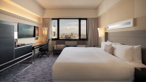 Une chambre d’hôtel avec un grand lit, un bureau avec une chaise, une télévision et une vue sur la ville par la fenêtre.