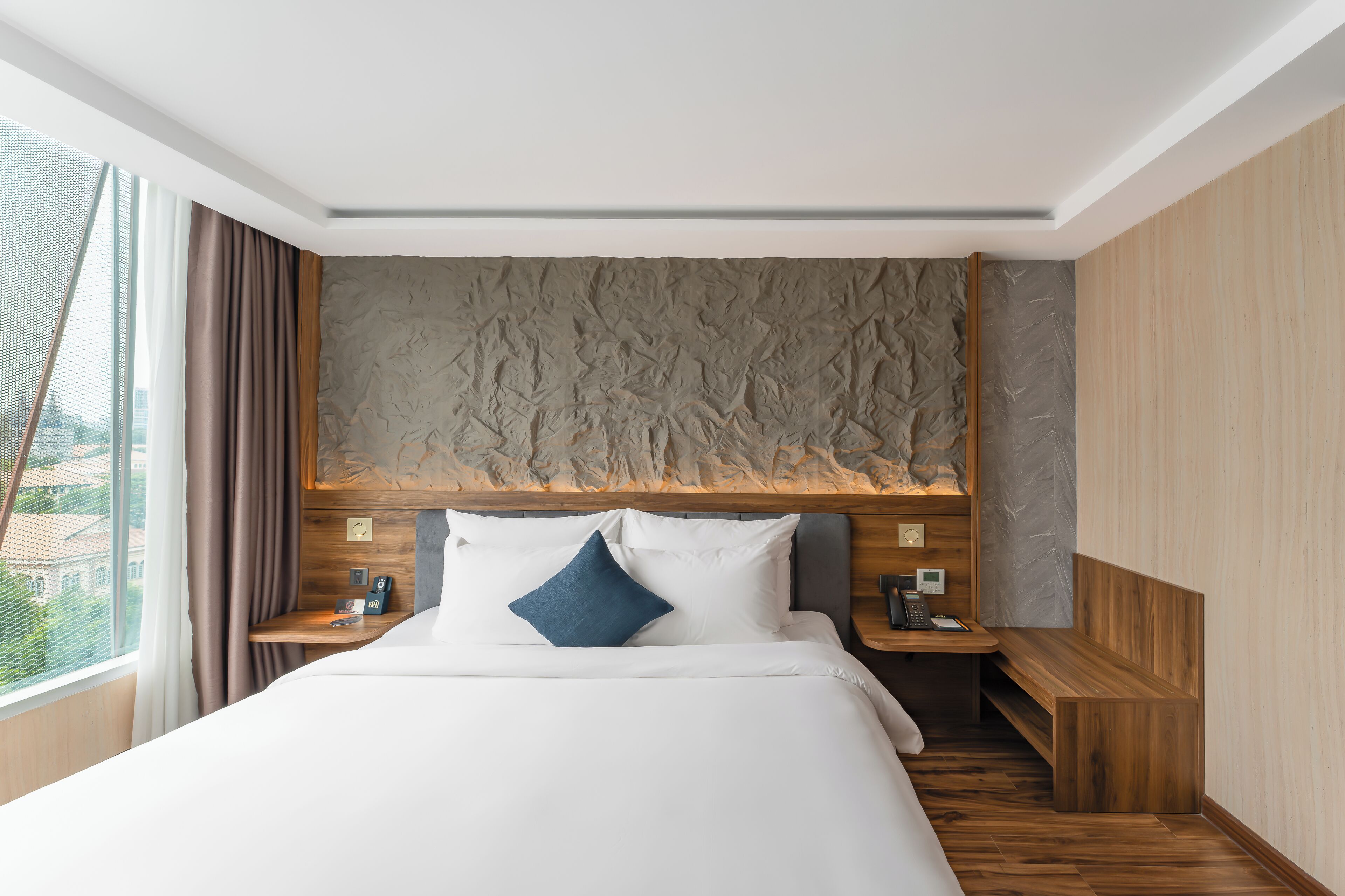 Ett modernt hotellrum med en stor säng, ett träbeklätt sänggavel och utsikt över staden genom ett stort fönster.