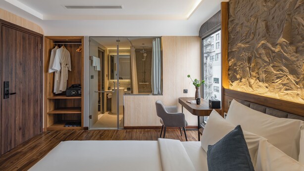Une chambre d’hôtel moderne avec une armoire en bois, un lit avec du linge de maison blanc, un bureau avec une chaise et une grande fenêtre donnant sur des immeubles.