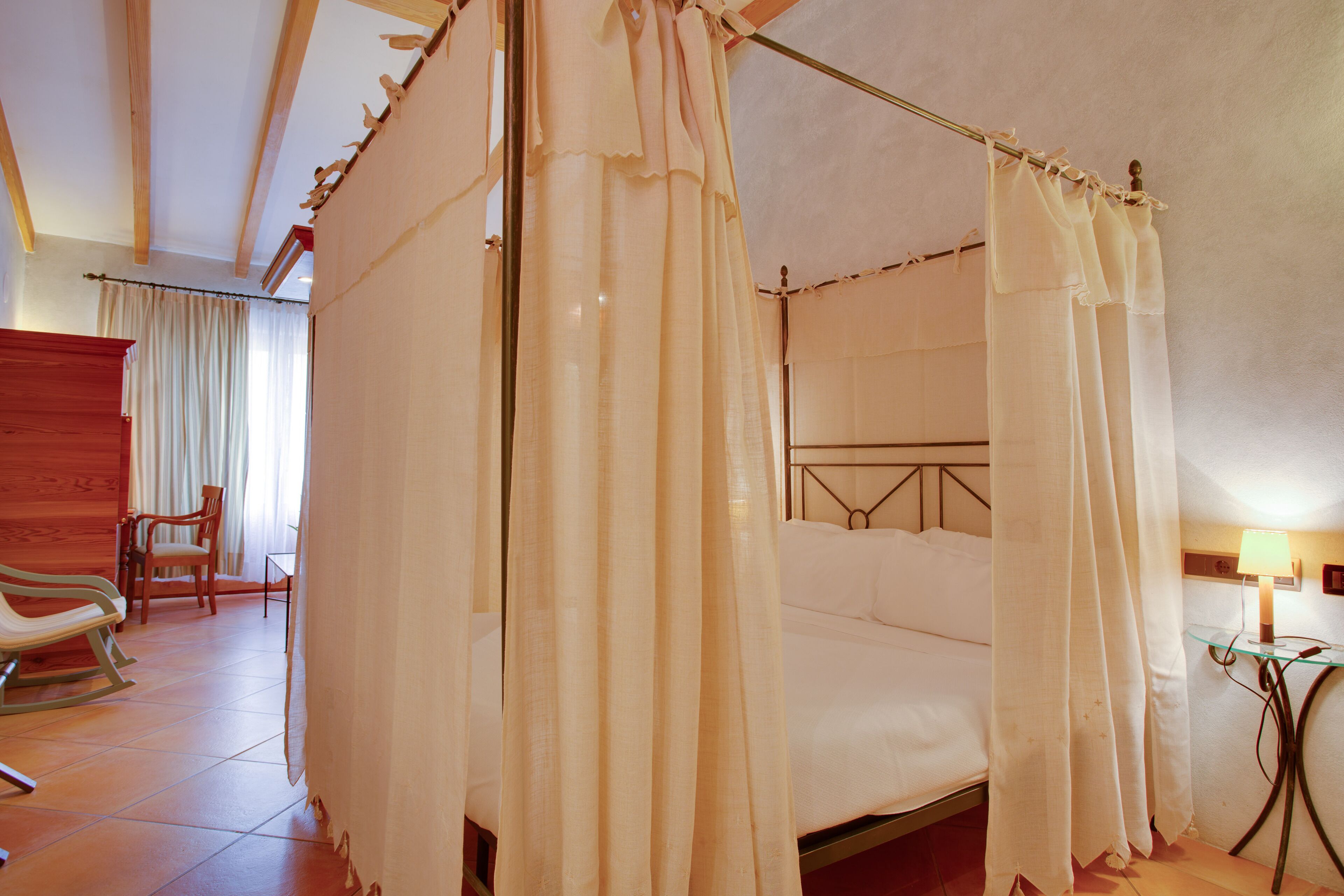 Una cama con dosel y pilares, una mecedora de madera, una mesita con una lámpara y una silla en una habitación de hotel.