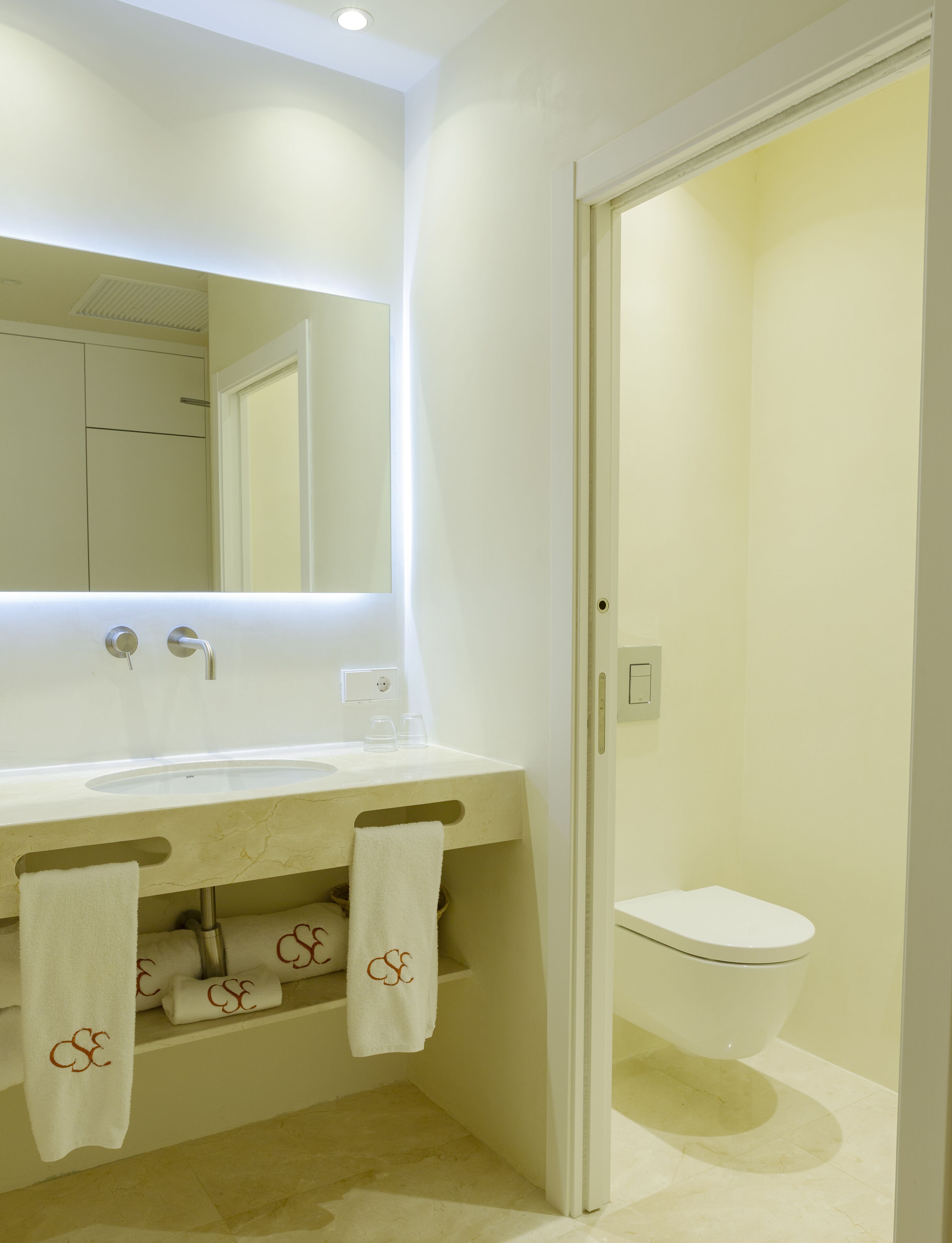 Une salle de bain moderne avec un lavabo, un miroir et des toilettes, le tout de couleur blanche. Des serviettes sont suspendues sur un porte-serviettes situé sous le lavabo.