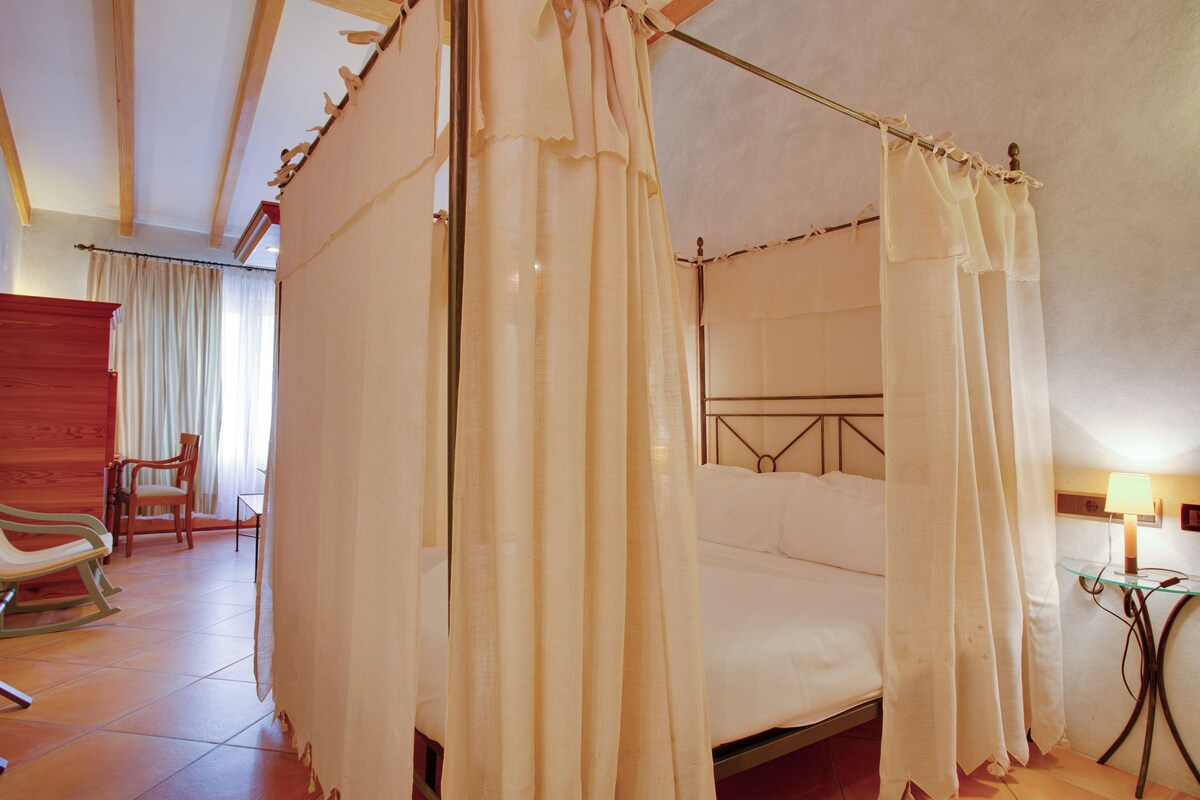 A four-poster bed with a canopy, a wooden rocking chair, a small table with a lamp, and a chair in a hotel room.