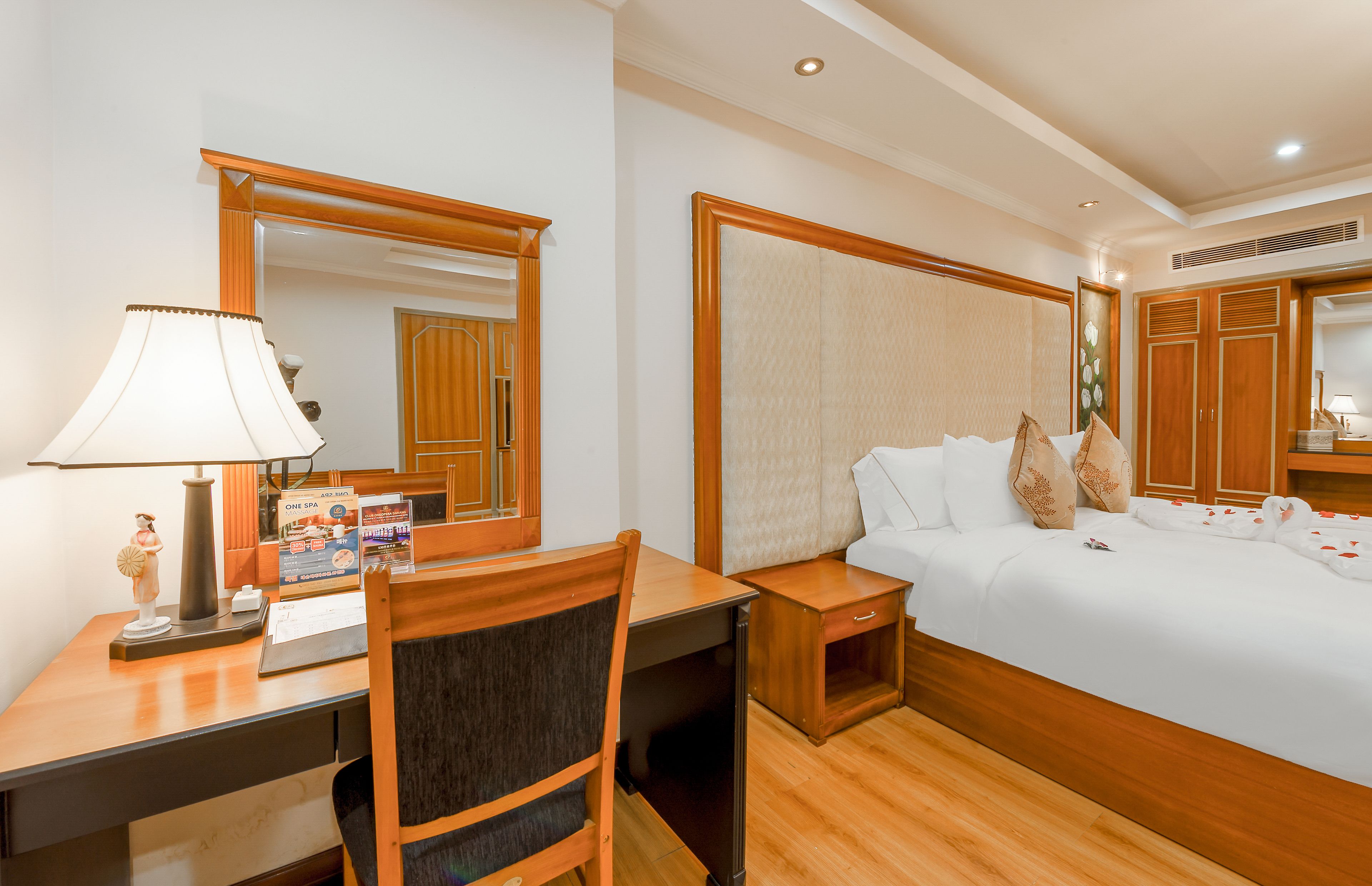 A hotel room with a bed, a desk with a lamp, a chair, a nightstand, and a mirror.