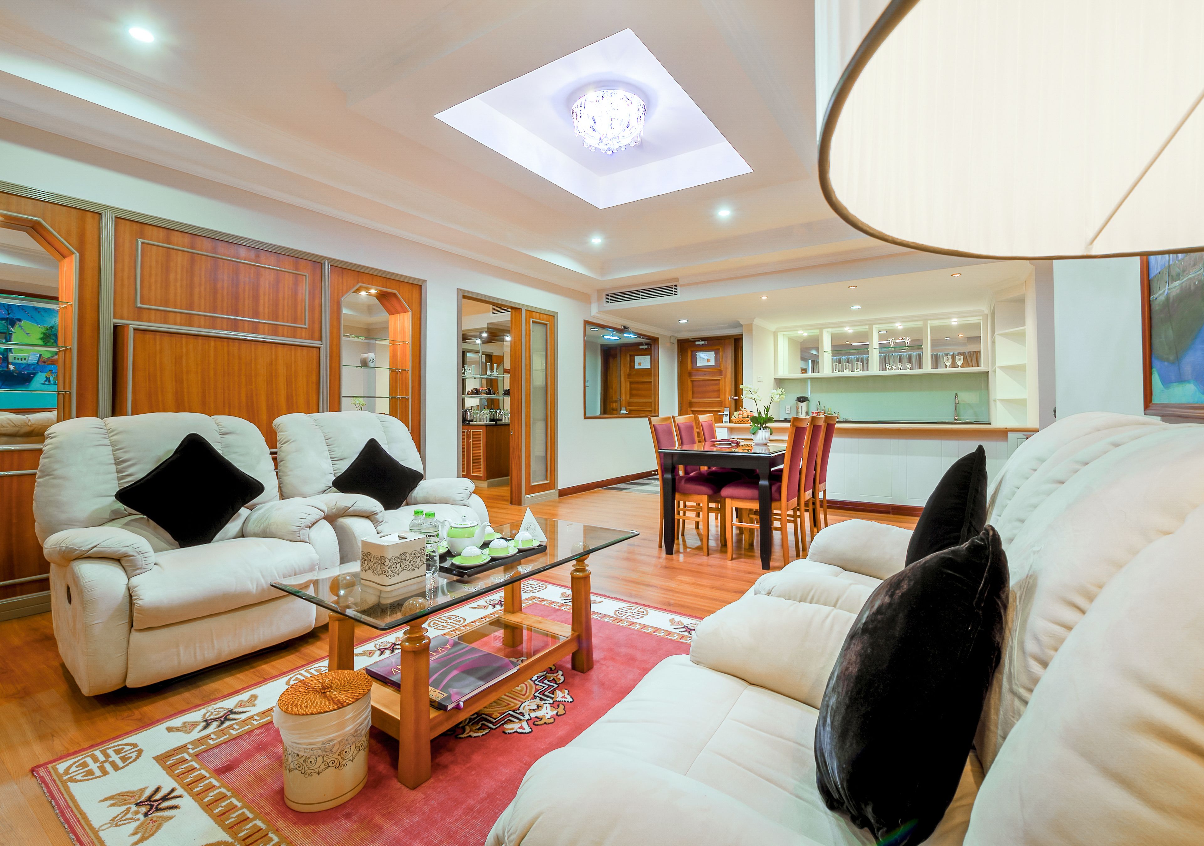A spacious living room with a glass coffee table, white sofas, and a dining area with wooden chairs.