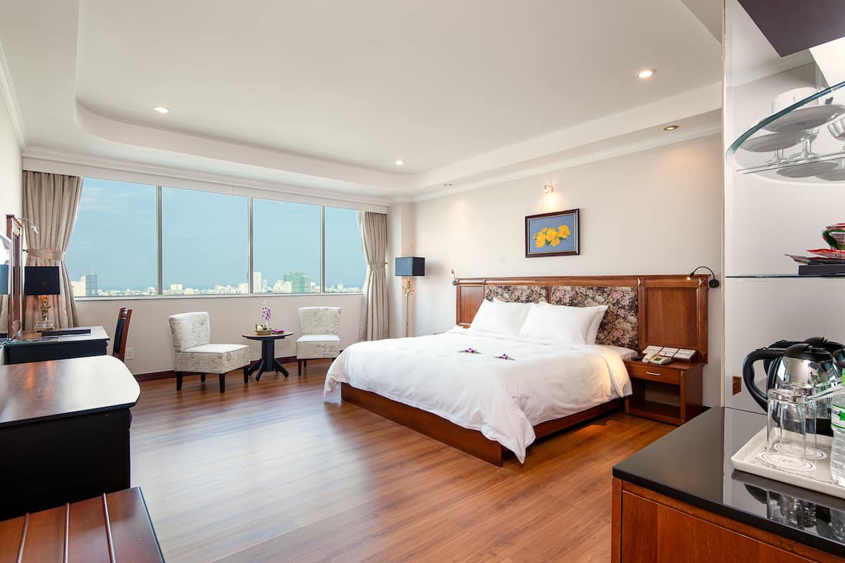 A hotel room with a large bed, a desk, a chair, a small table, and a view of the city.