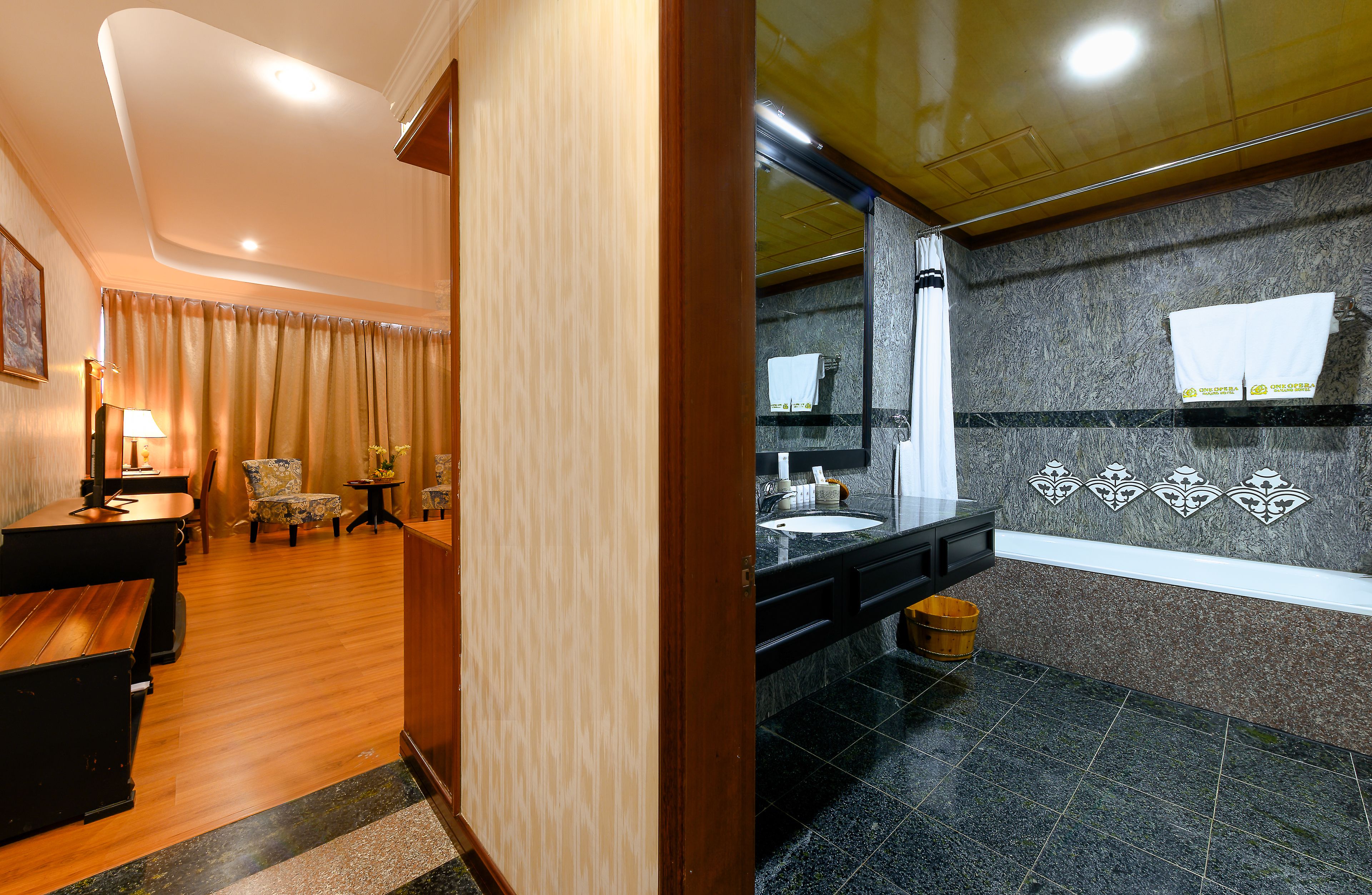 A hotel room with a wooden floor, a sitting area with chairs, a desk, and a large window with curtains.