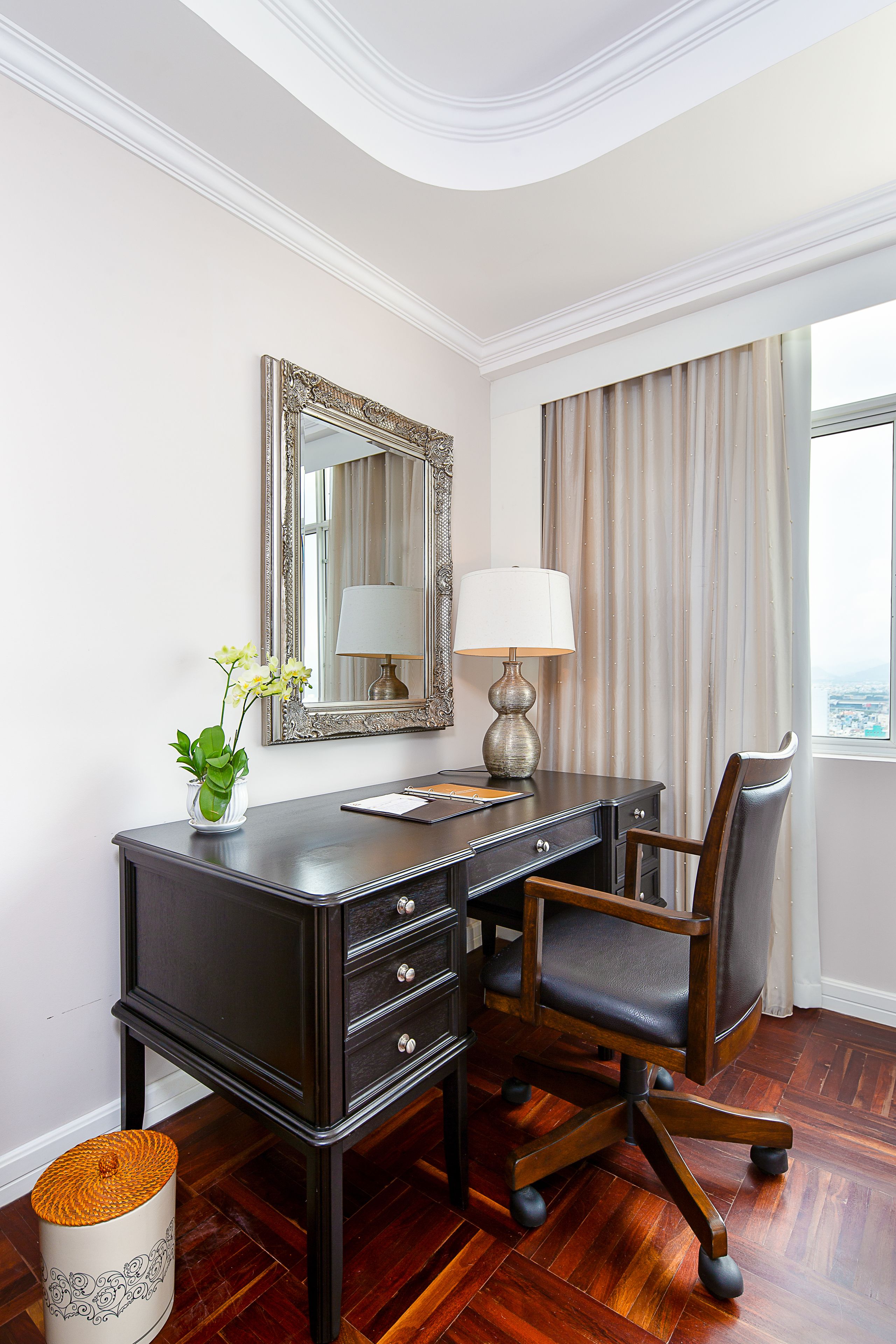 A hotel room with a dark wooden desk, a chair, a mirror, a lamp, and a window with curtains.