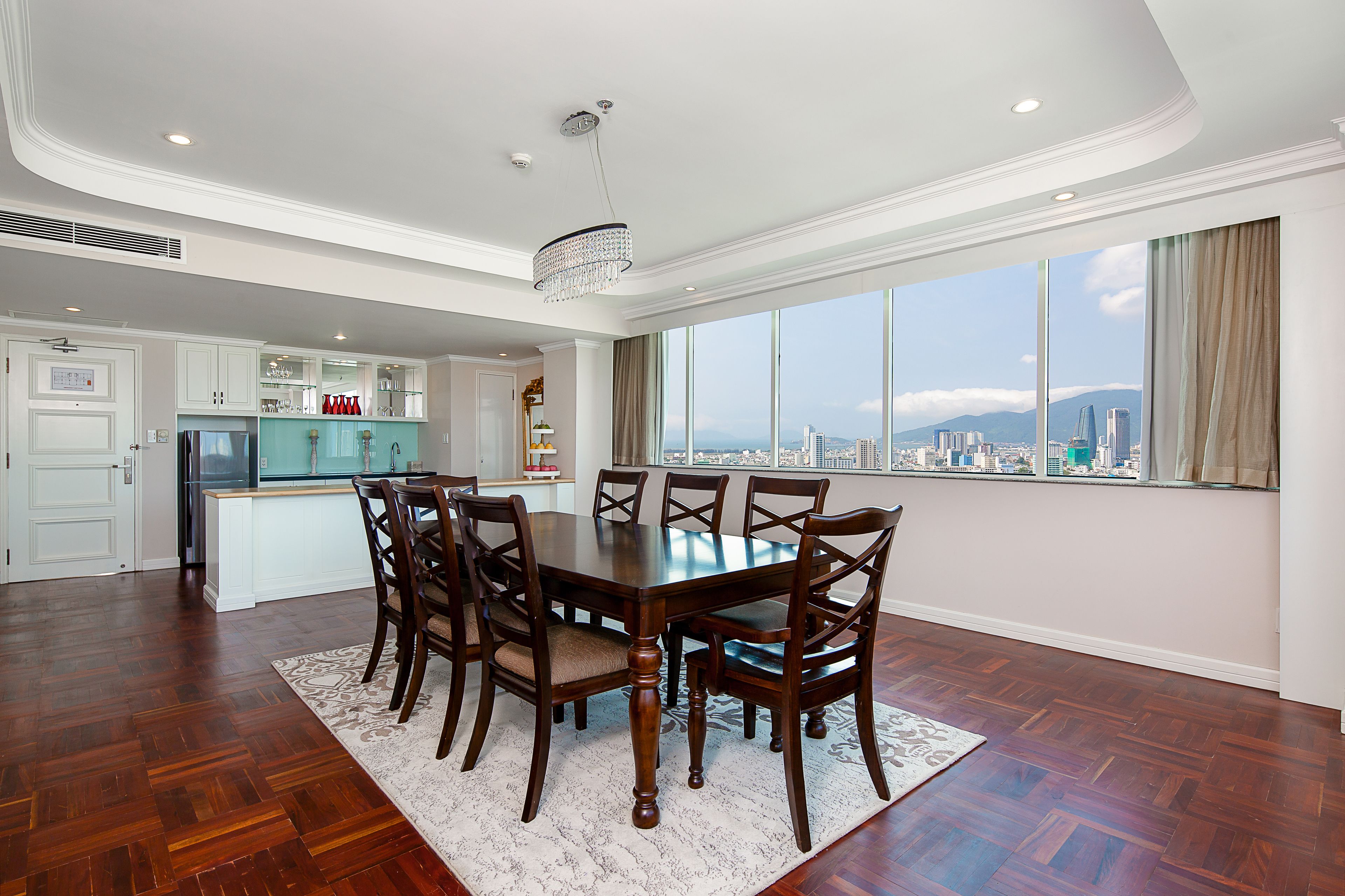 A spacious dining area with a large wooden table and chairs, a chandelier, and a view of the city through large windows.