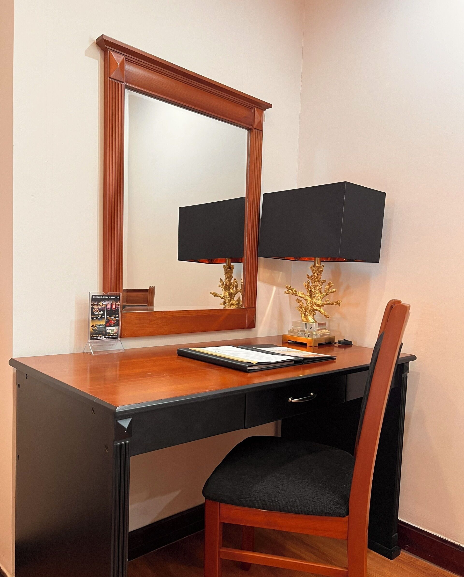 A hotel room with a wooden desk, a chair, a mirror, a lamp, and a small decorative plant.