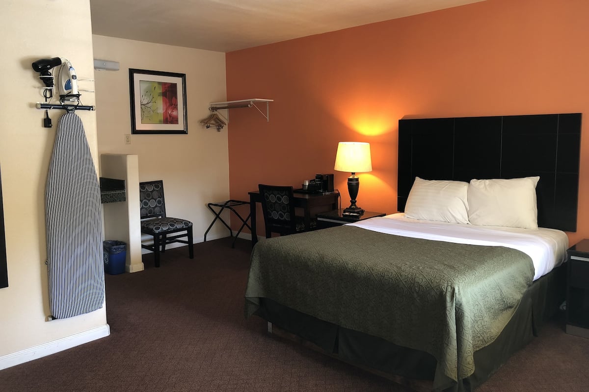 A hotel room with a bed, a desk, a chair, a lamp, and a coat rack.