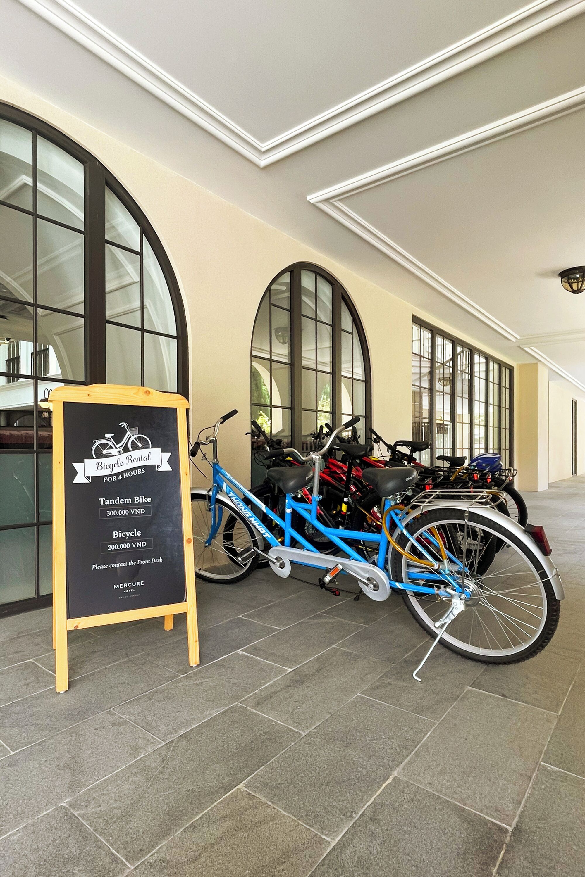 Un panneau indiquant les tarifs et les coordonnées pour la location de vélos, un vélo tandem bleu et plusieurs autres vélos garés dans un espace intérieur doté de grandes fenêtres et d’un sol carrelé.