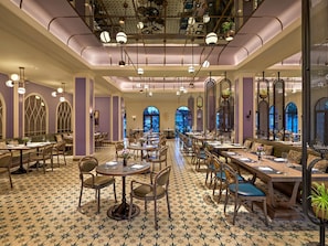 A spacious dining area with multiple tables and chairs, large windows, and a high ceiling with hanging lights.