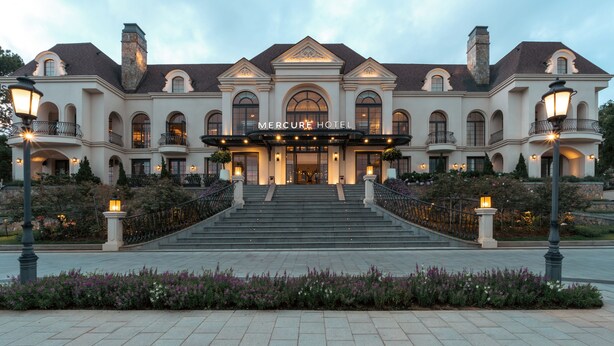 A grand hotel entrance with a symmetrical facade, large arched windows, and a central entrance flanked by steps. - Grand Mercure Dalat (Lam Vien - Da Lat)