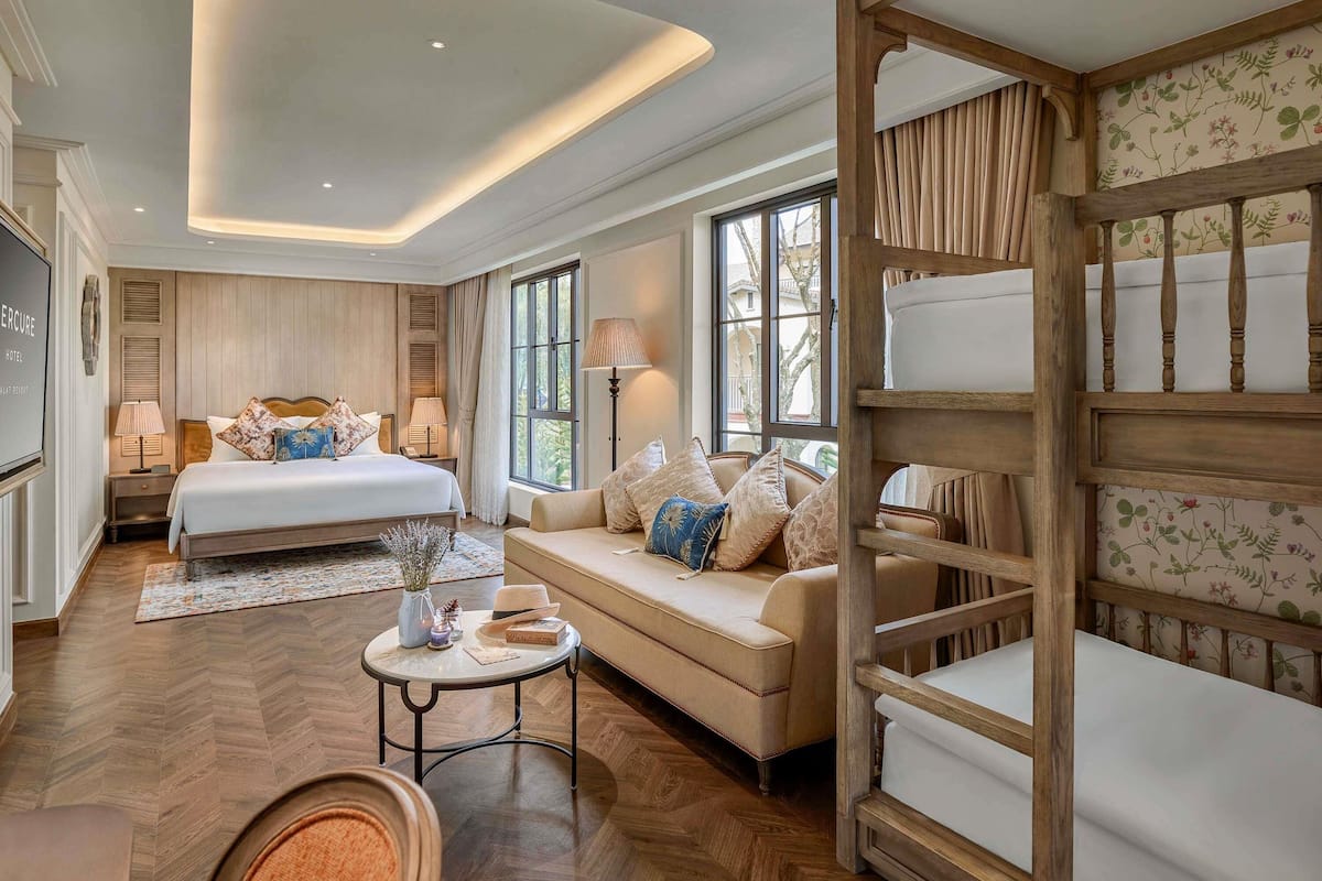 Habitación de hotel con cama, sofá, banco de madera, mesa redonda y una ventana grande.