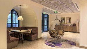 A spacious lobby with a high ceiling, a seating area with a table and cushioned benches, a hanging chair, and a decorative rug. - Grand Mercure Dalat (Lam Vien - Da Lat)