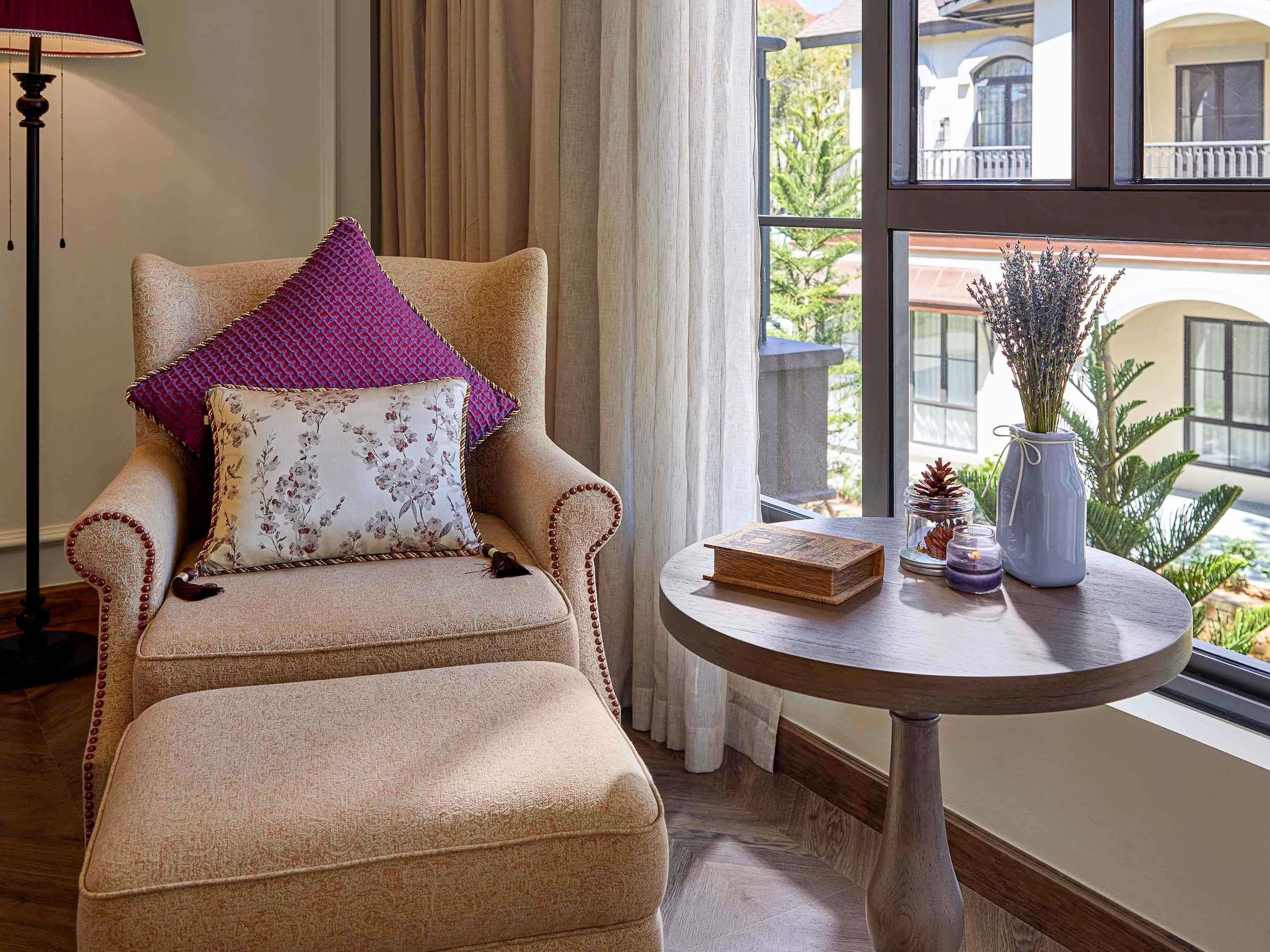 A beige armchair with floral and patterned cushions, a small round table with a book and decorative items, and a view of a building through a window.