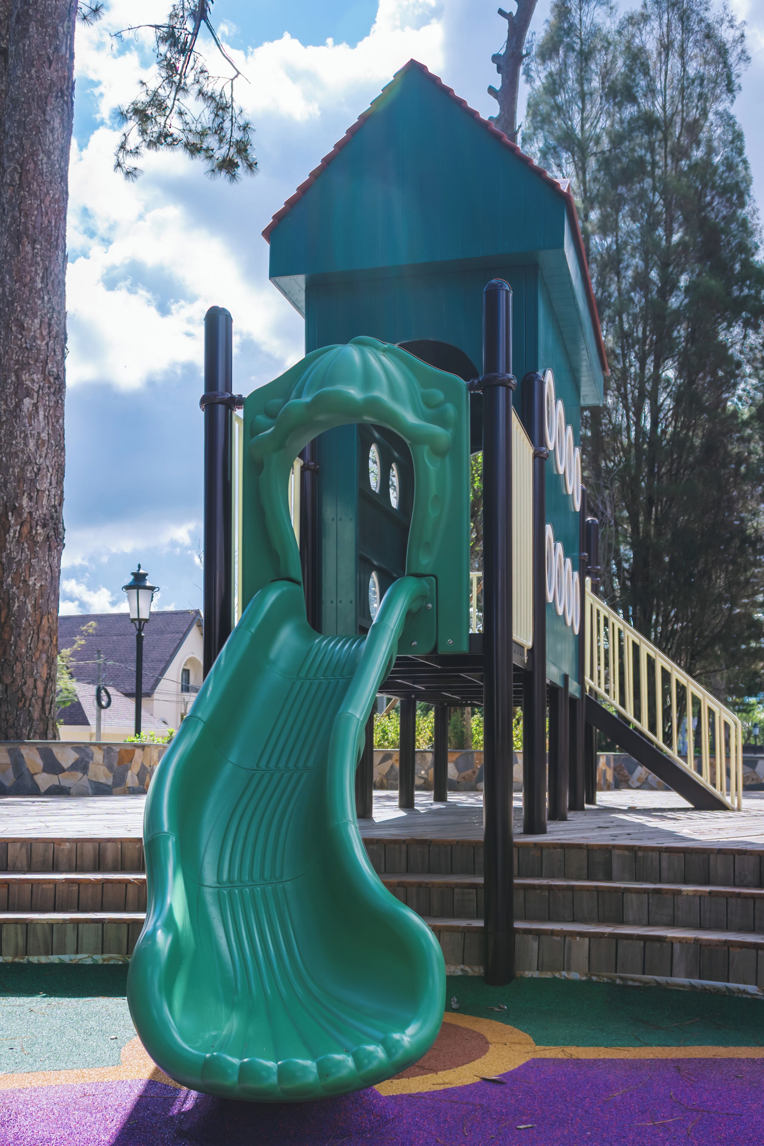 Un parque infantil con un tobogán verde, una estructura para escalar y un pequeño edificio con techo rojo.